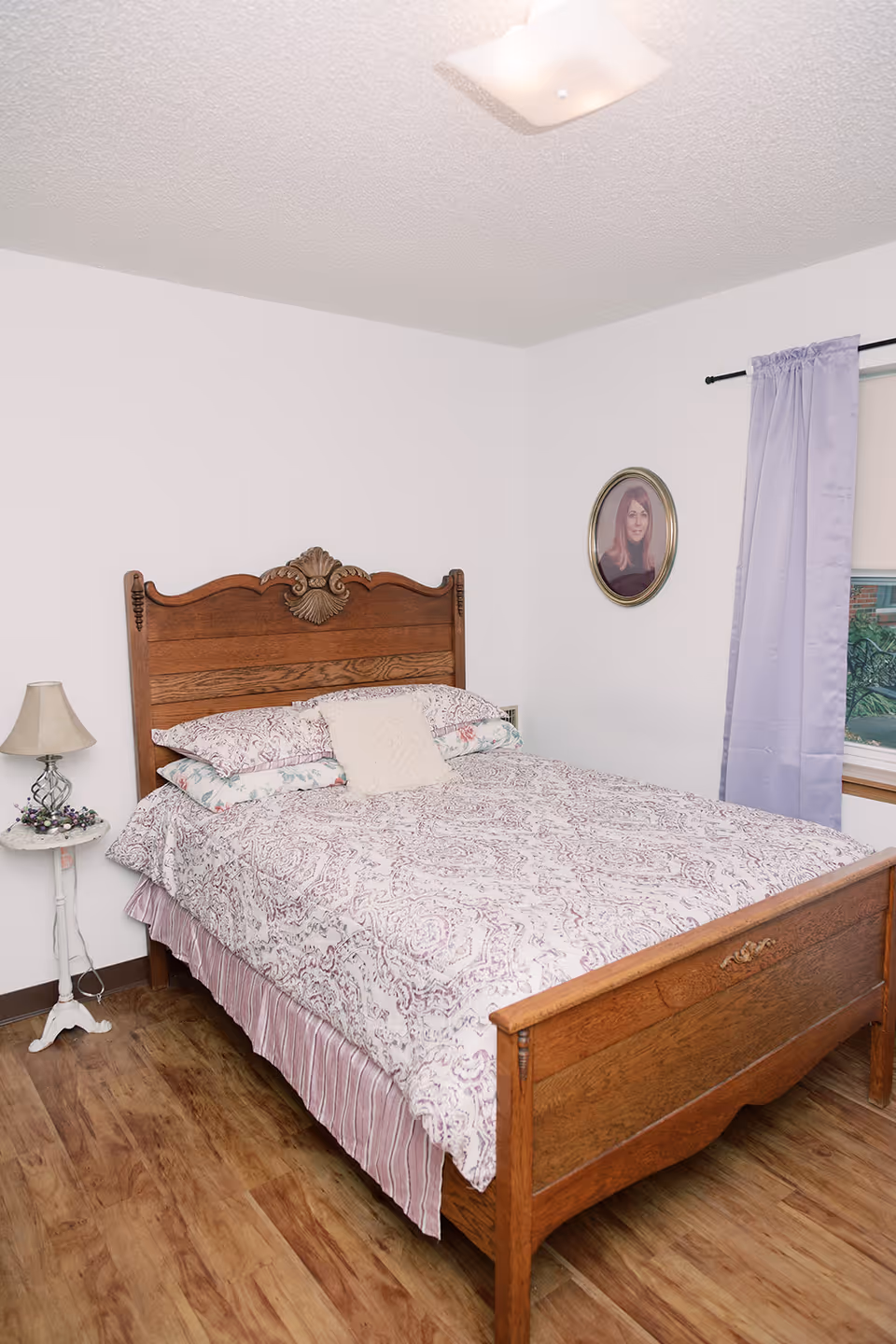 A bedroom with a wooden bed frame featuring an ornate headboard and footboard. The bed is made with a patterned bedspread and multiple pillows. To the left of the bed is a small white side table with a lamp and decorative items. On the wall above the bed is a framed oval portrait of a woman. A window with light purple curtains is visible on the right side of the room. The floor is wooden and the walls are painted white.