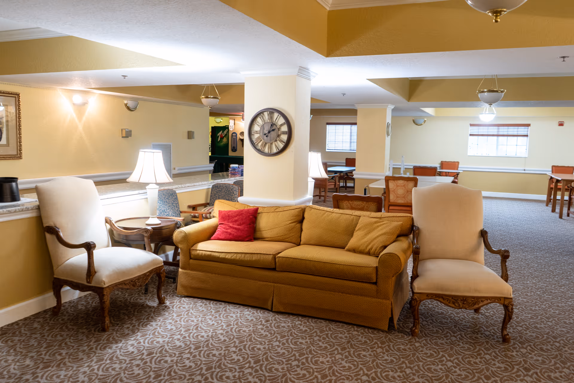 A cozy senior living common area with a mustard yellow sofa adorned with a red pillow, flanked by two beige upholstered armchairs with wooden frames. The room features patterned carpet, warm yellow walls, a large round wall clock on a column, and several tables and chairs in the background near windows with blinds. Soft lighting is provided by table lamps and ceiling fixtures.