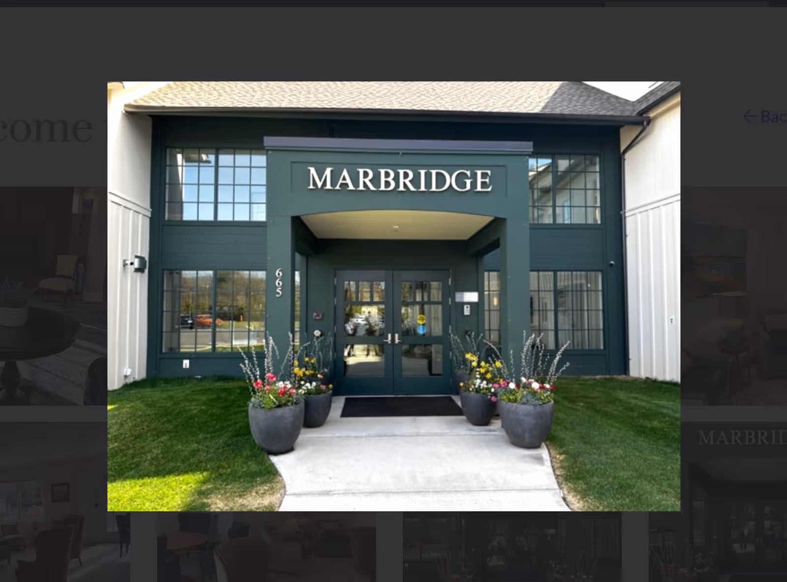 Front entrance of Marbridge Assisted Living & Memory Care facility with green double doors, large windows on either side, and the name MARBRIDGE displayed above the entrance. There are five large flower pots with colorful flowers arranged symmetrically along the walkway leading to the doors.