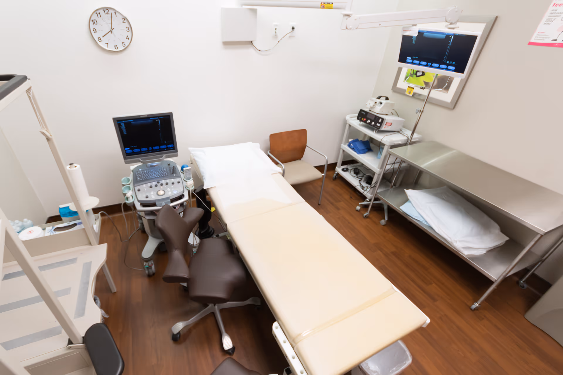 A medical procedure room with an examination bed, monitors, and stainless steel shelving.