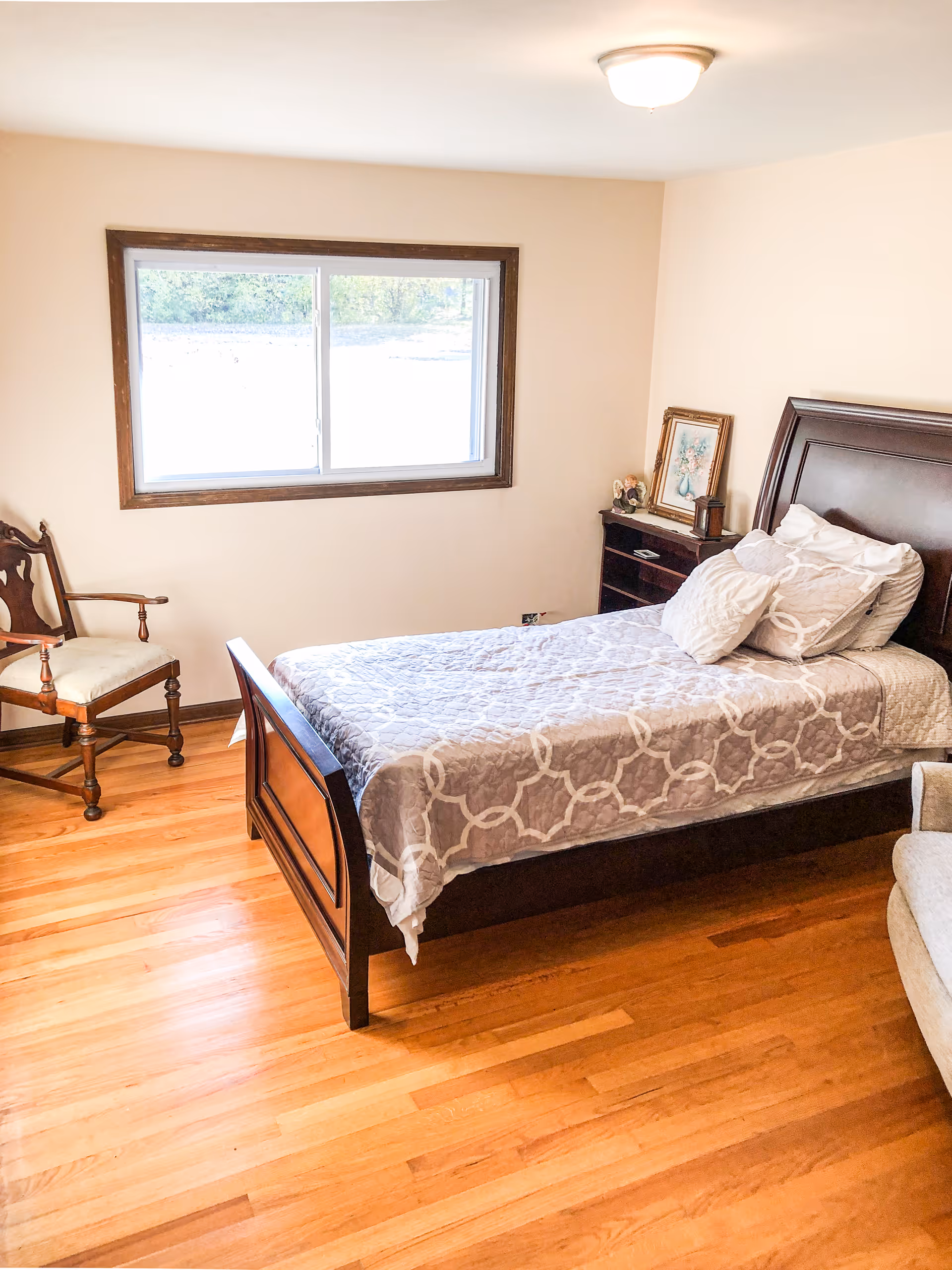 A bright bedroom with a wooden bed frame and a neatly made bed with patterned bedding. There is a wooden chair with a cushioned seat near the window, a small dark wooden nightstand with a framed picture and decorative items, and a light fixture on the ceiling. The room has hardwood floors and a large window letting in natural light.