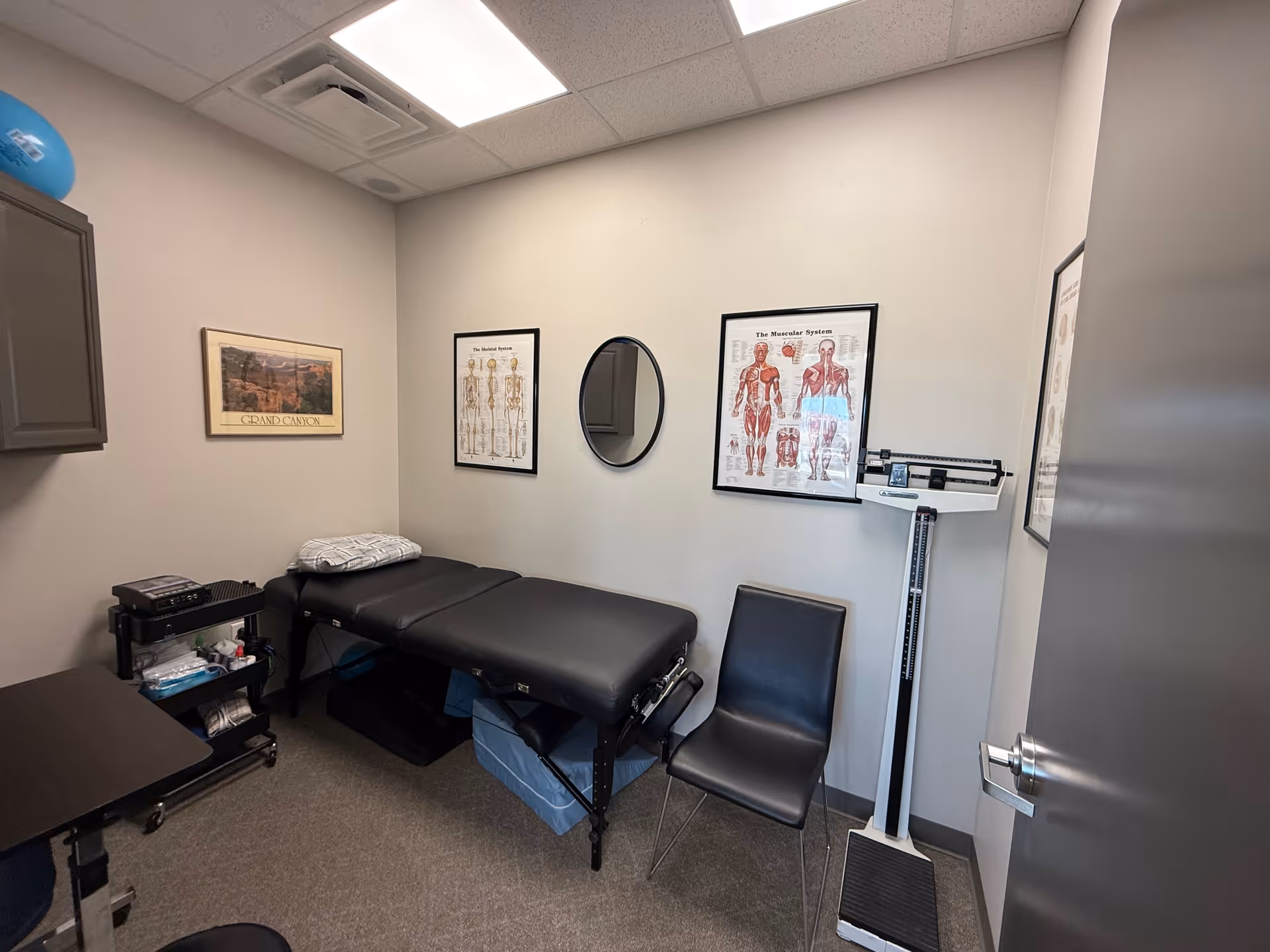 Therapy room with a black treatment table, medical charts, a mirror, a scale, and chairs.