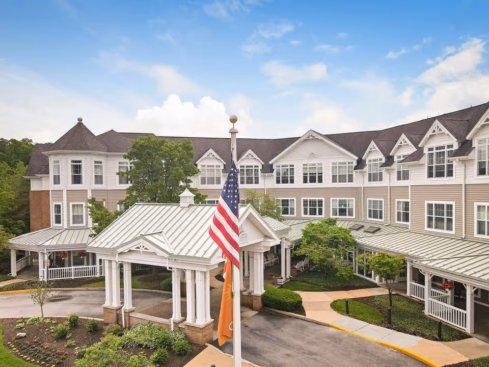 Front entrance of a multi-story senior living building with a covered porte-cochere, landscaped driveway, and an American flag.