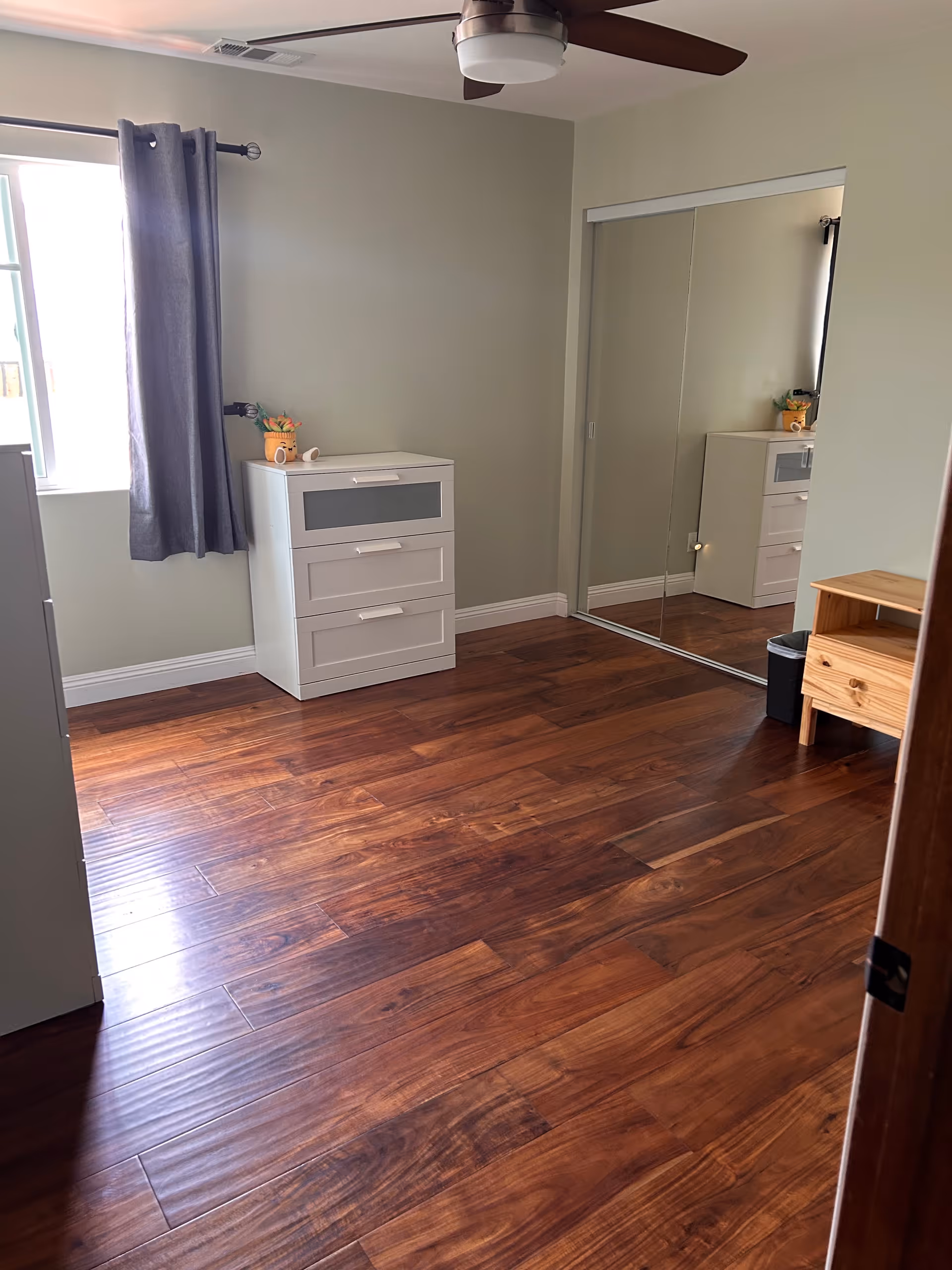 A bedroom with wooden flooring, dressers, a window with gray curtains, and a mirrored closet.