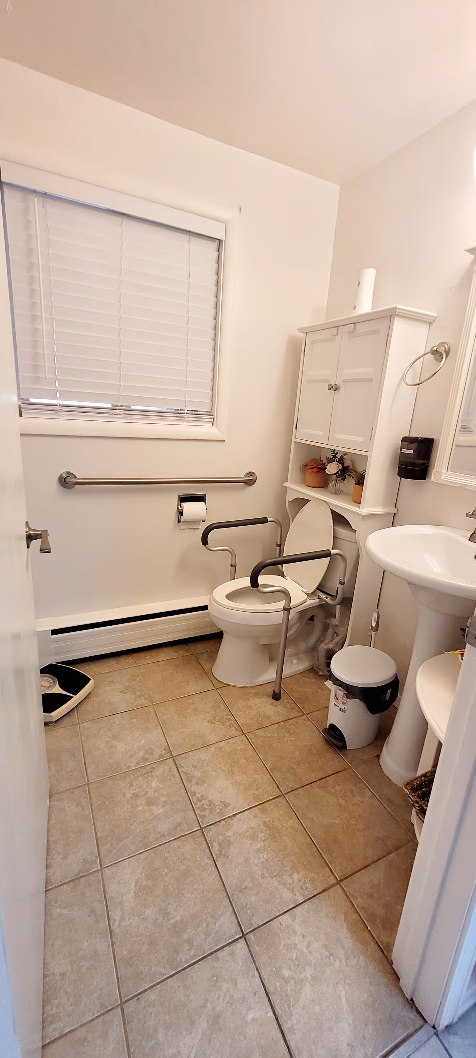 Accessible bathroom with toilet, grab bars, sink, cabinet, window blinds, and tiled floor.