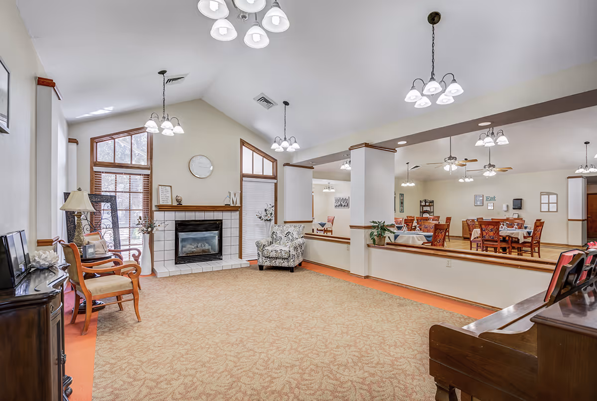 Spacious senior living lounge with fireplace, seating, a piano, and a dining area in the background.