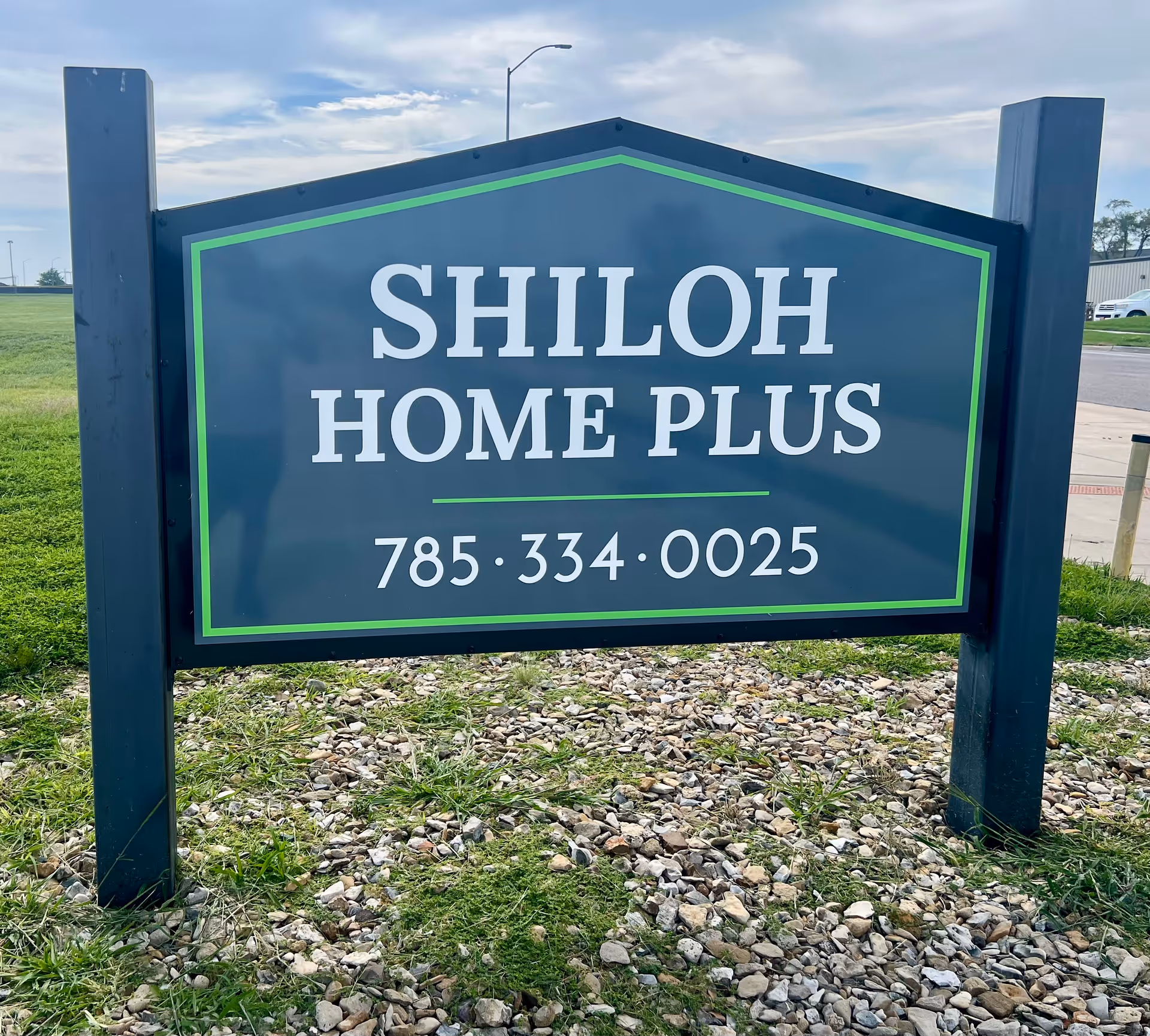 Blue-green sign reading SHILOH HOME PLUS with phone number stands outdoors between posts.