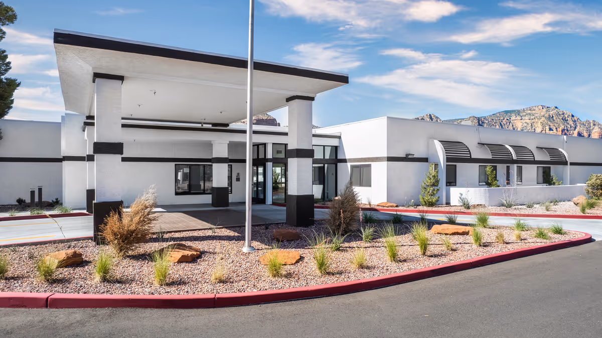 Exterior view of a modern single-story building with white walls and black trim, featuring a covered entrance supported by columns. The surrounding landscape includes desert plants, rocks, and a paved driveway with a red curb. Mountains and a partly cloudy sky are visible in the background.