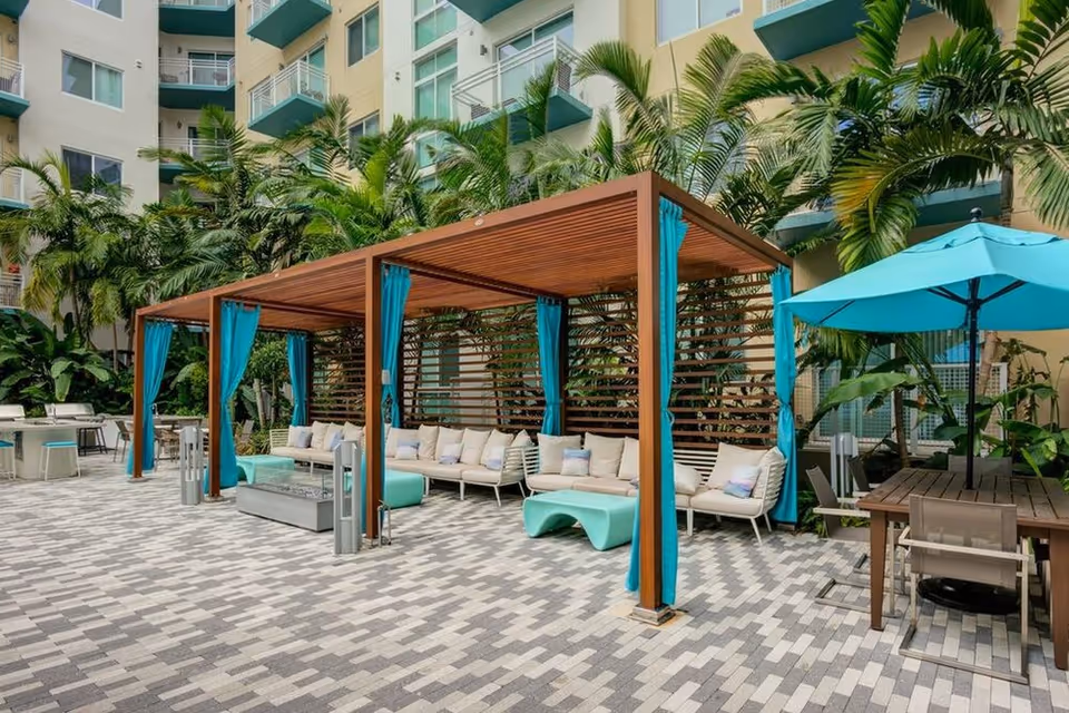 Outdoor courtyard with wooden cabanas draped in teal curtains, lounge seating, umbrellas, and palm trees in front of a multi-story building.