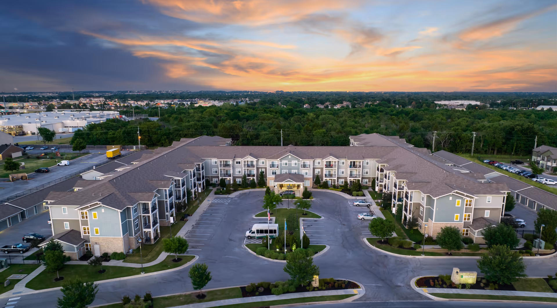 Aerial view of The Cedars at Woodland Hills senior living facility at sunset, showing a large U-shaped building surrounded by parking lots, trees, and greenery with a colorful sky in the background.