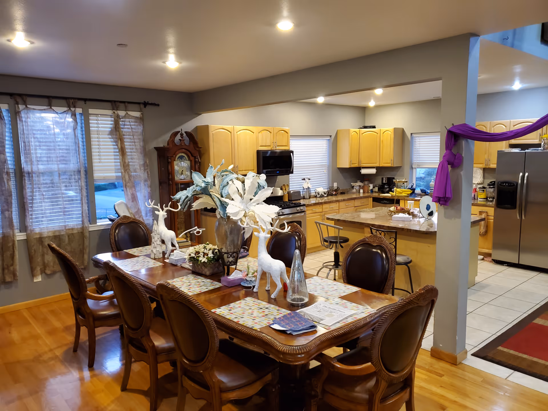 A spacious dining area with a large wooden dining table surrounded by eight ornate chairs. The table is decorated with placemats, white decorative reindeer figures, a large vase with white flowers, and other small decorative items. Behind the dining area is an open kitchen with light wood cabinets, a stainless steel refrigerator, a stove, and a kitchen island with bar stools. The room has wooden flooring in the dining area and tiled flooring in the kitchen. Windows with sheer curtains allow natural light into the space.
