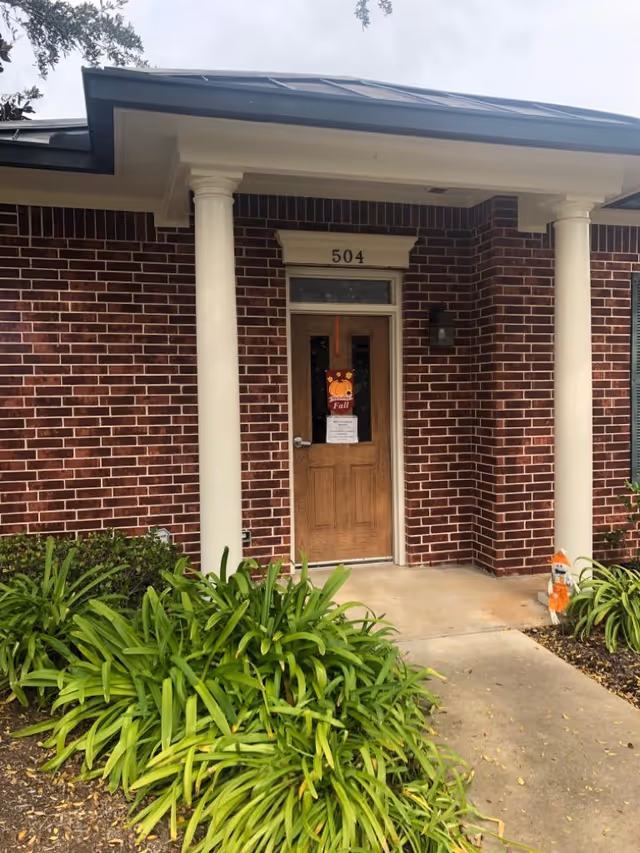 Brick building entrance with two white columns, door, and landscaping.