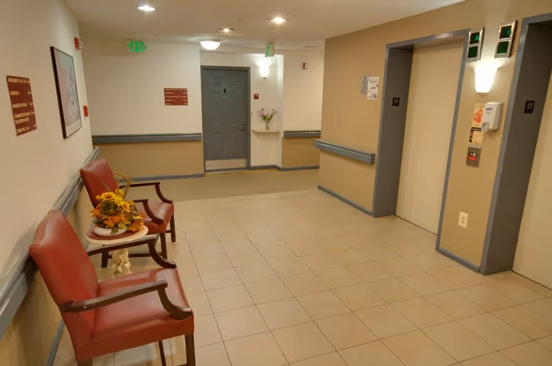 A tiled interior lobby with two red chairs and a small table with flowers, a nearby door, and two elevator doors.