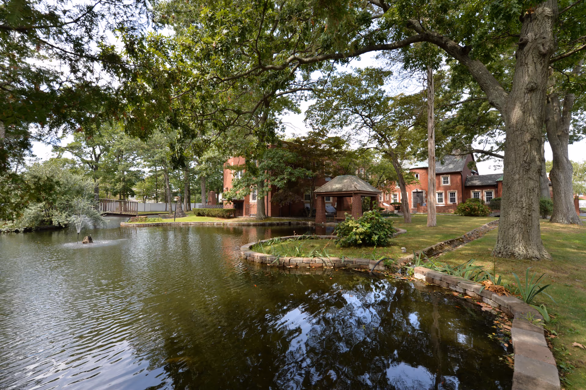 Birchwood Suites exterior: brick buildings, pond, gazebo, and shaded lawn.