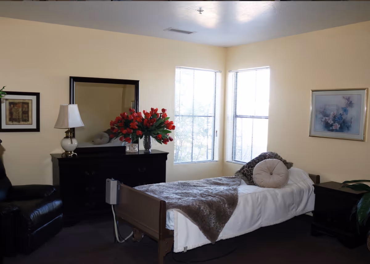 Sunlit bedroom with single bed, dresser, mirror, lamp, and flower arrangement.