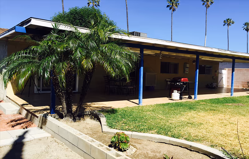 Outdoor patio area of a facility with a covered seating area supported by blue pillars, a barbecue grill, a small palm tree, and a grassy lawn under a clear blue sky.