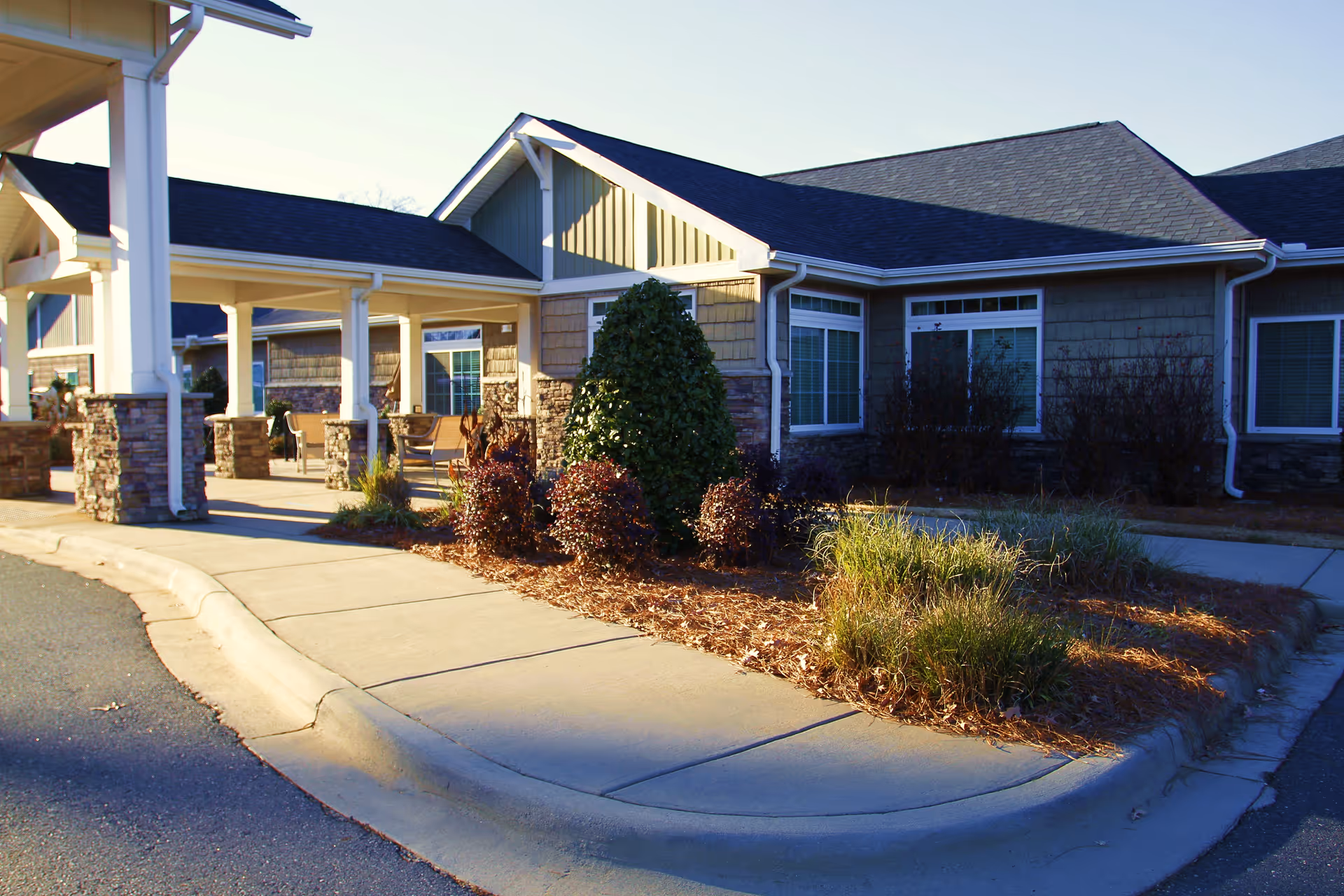 Exterior view of a senior living facility with a covered walkway supported by white columns with stone bases, landscaped bushes, and a paved sidewalk under clear daylight.