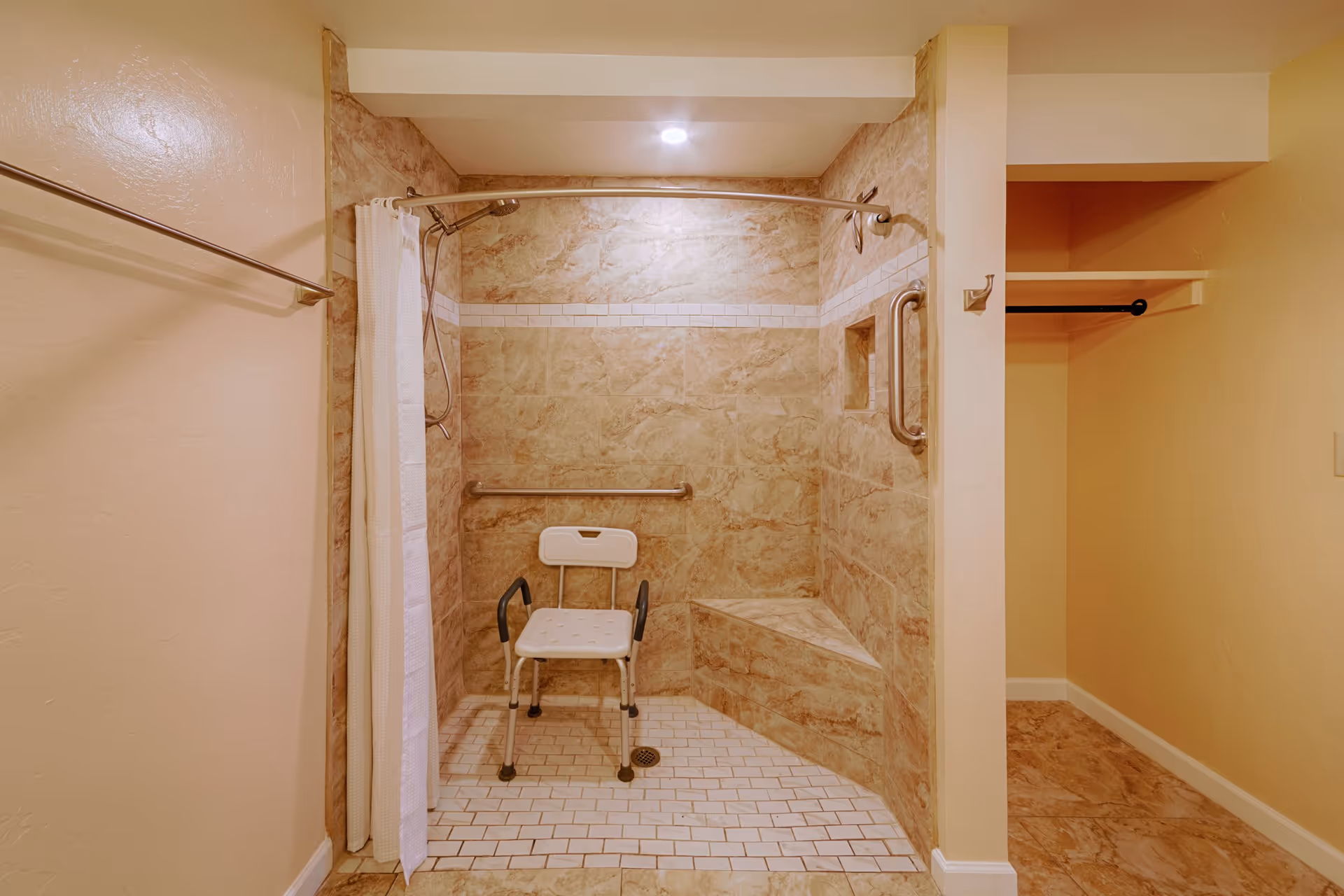 A bathroom with a walk-in shower featuring beige marble tiles, a white shower chair with armrests, a grab bar on the wall, a built-in corner seat, and a white shower curtain on a curved rod. Adjacent to the shower is an open closet area with a hanging rod.