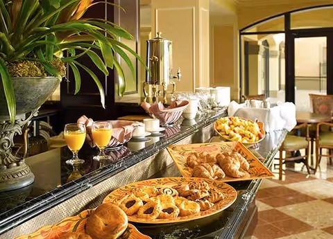 Buffet table with pastries, fresh fruit, glasses of orange juice and a coffee urn in a dining area.