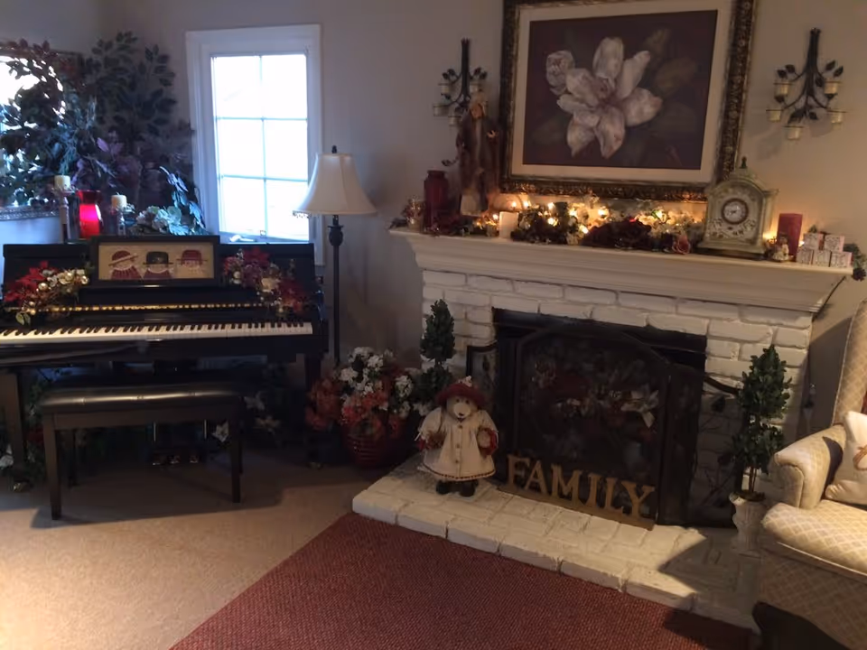 Cozy living room with a decorated white brick fireplace, piano, floor lamp, floral artwork, and a 'FAMILY' sign on the hearth.