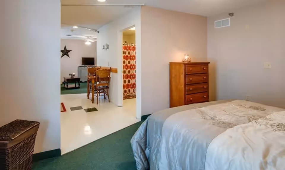 Bedroom with a made bed and wooden dresser opening into a dining/living area with a table, chairs and a TV.