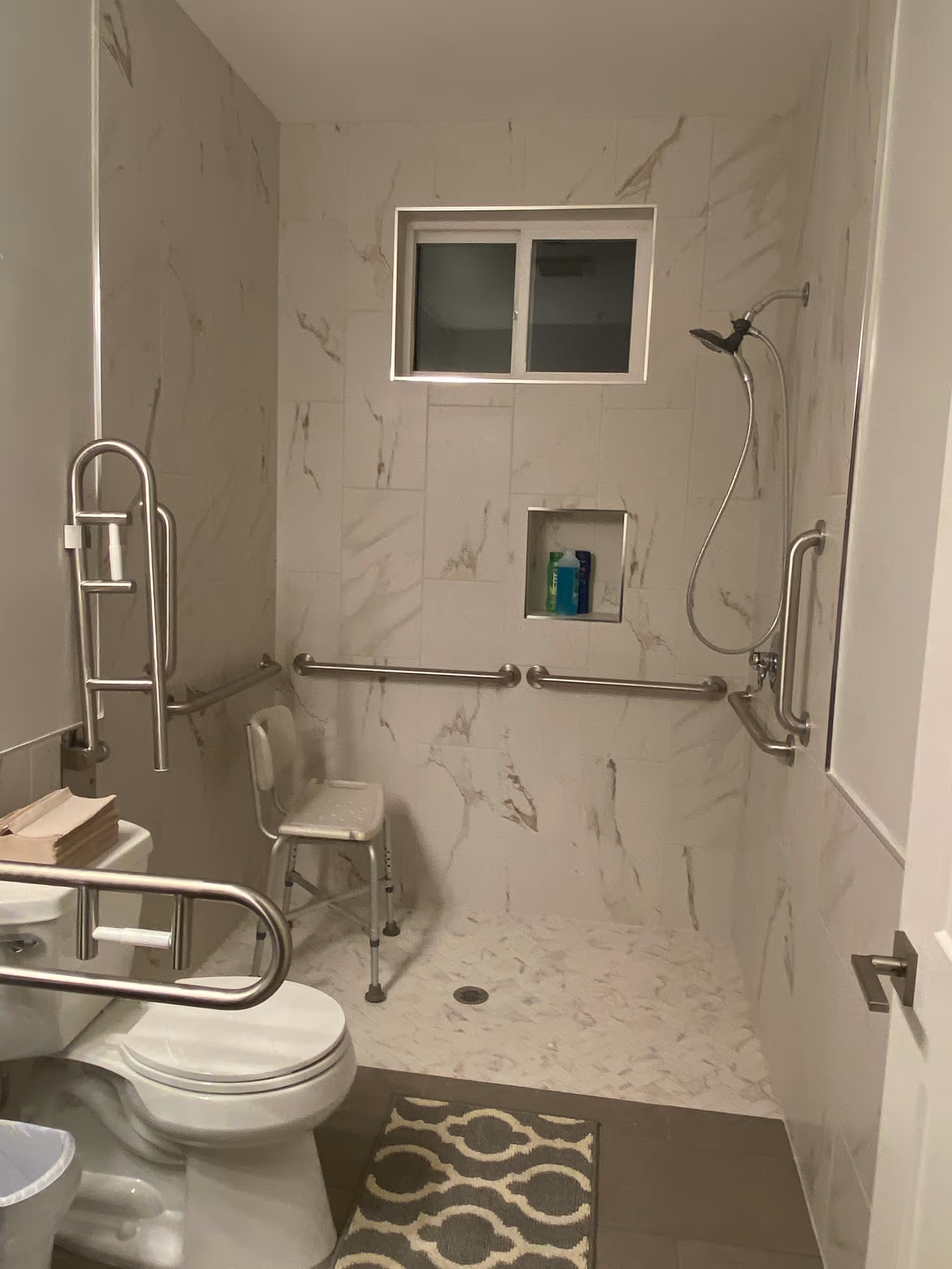 Accessible marble-tiled bathroom with toilet, shower, grab bars, and a chair.