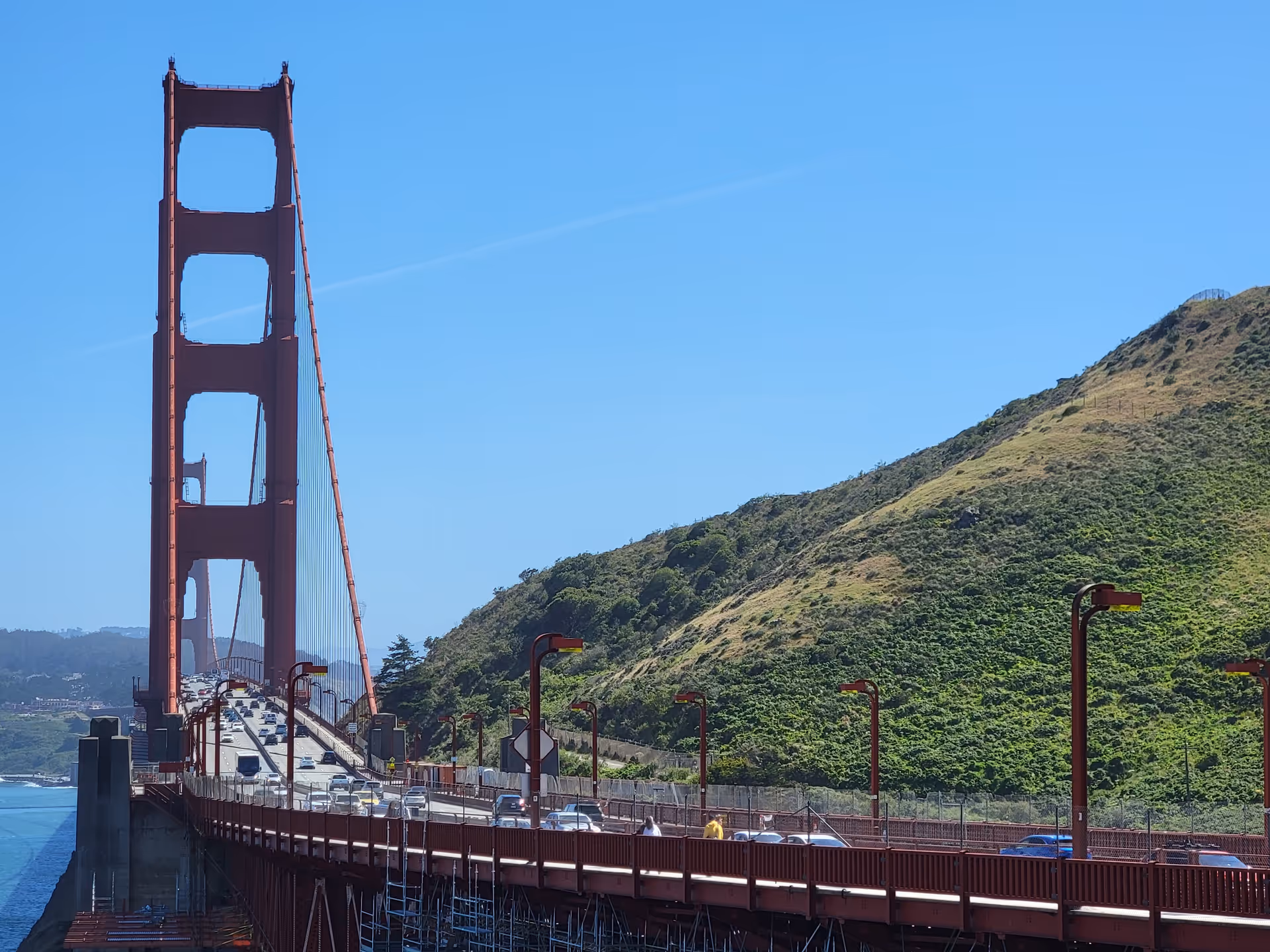 The Golden Gate Bridge stretches across a blue sky over green hills.