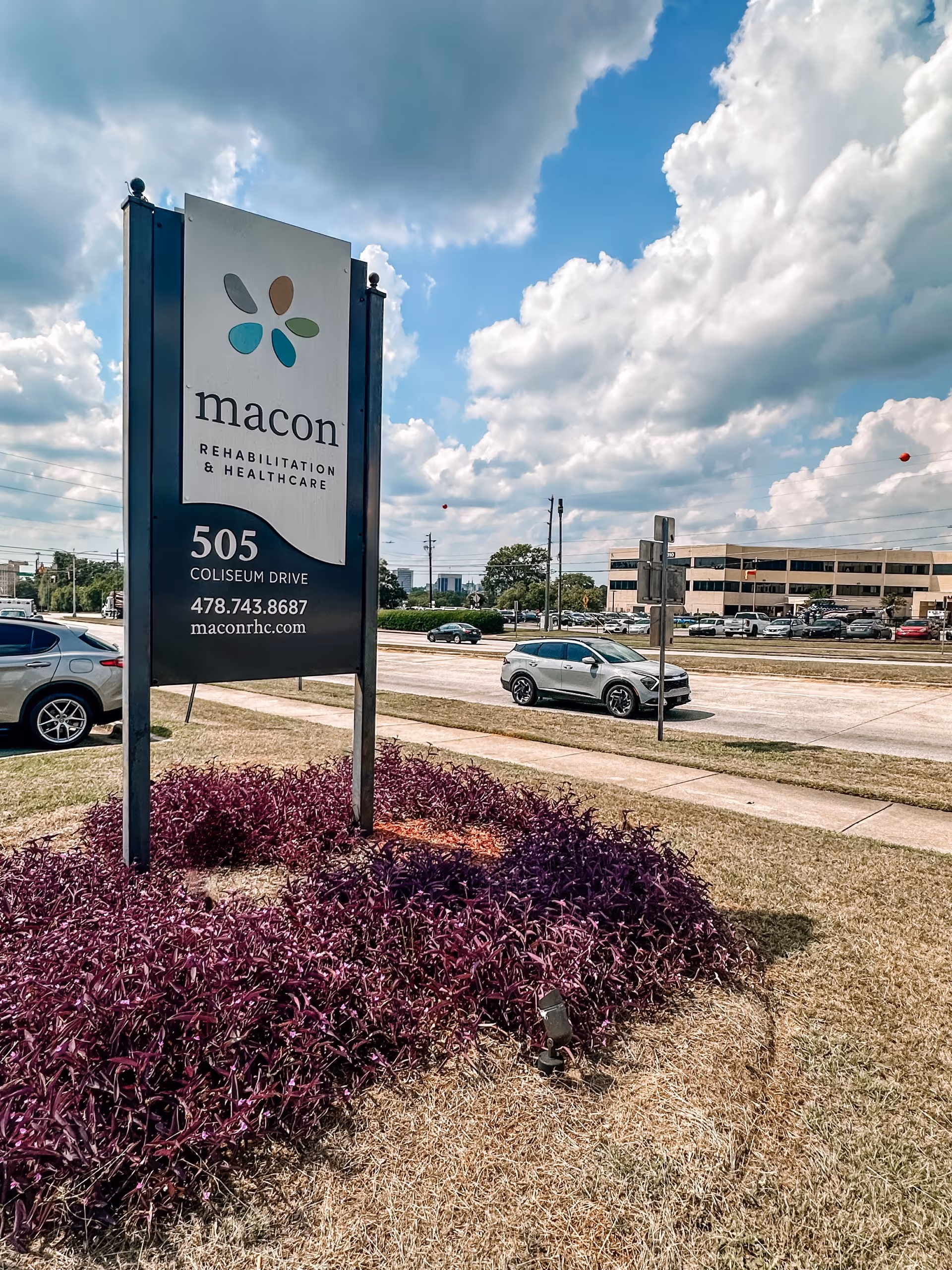 Outdoor photo of a Macon Rehabilitation & Healthcare roadside sign with purple landscaping, cars, and a building in the background under a partly cloudy sky.