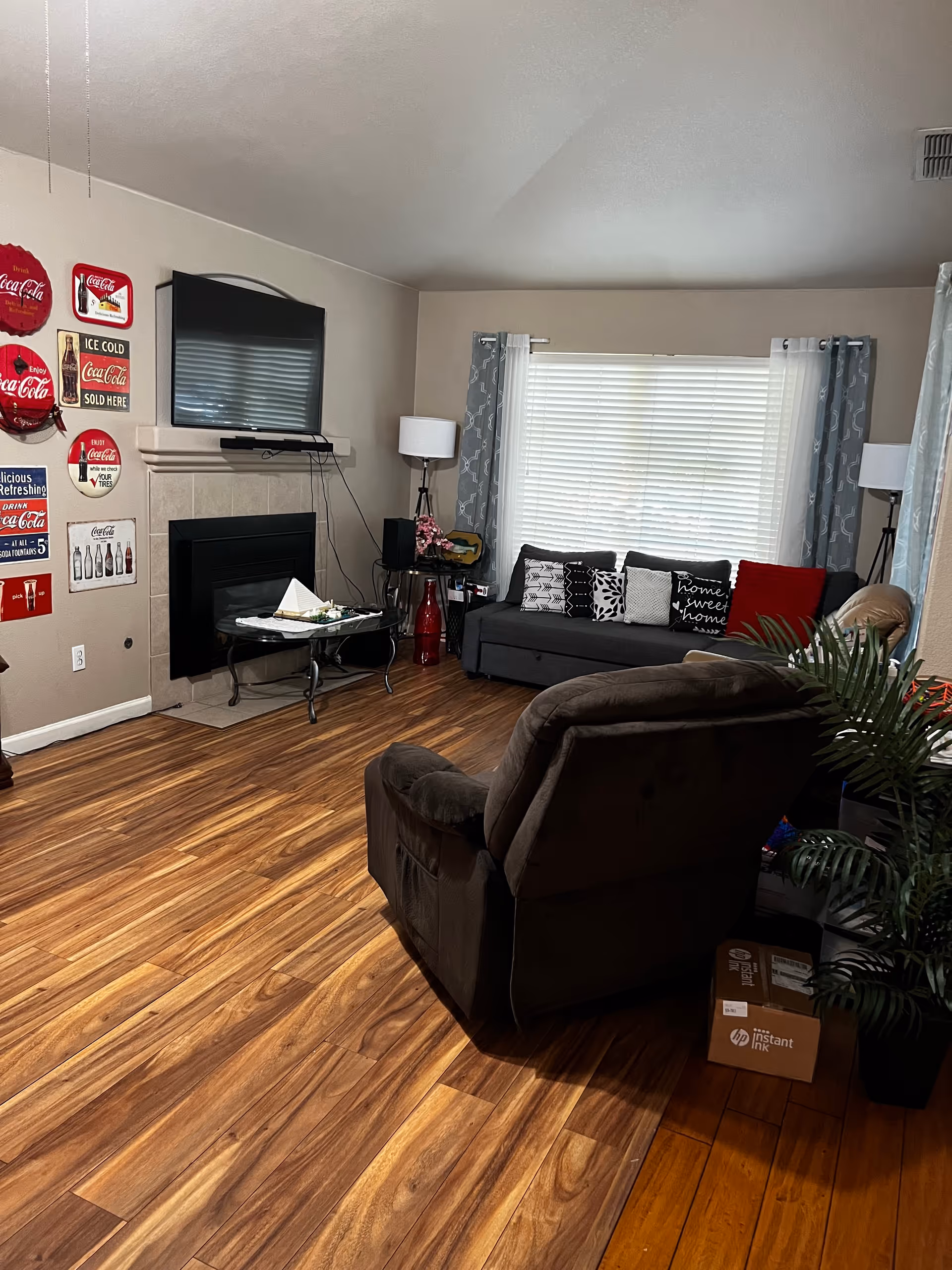 A cozy living room with wooden flooring, a dark gray sofa adorned with various patterned cushions including one that says 'home sweet home', a brown recliner chair, a glass coffee table with a decorative item, a fireplace with a mounted TV above it, and several Coca-Cola themed wall decorations on the left wall. There are two floor lamps on either side of the sofa and a large window with white blinds and gray patterned curtains.