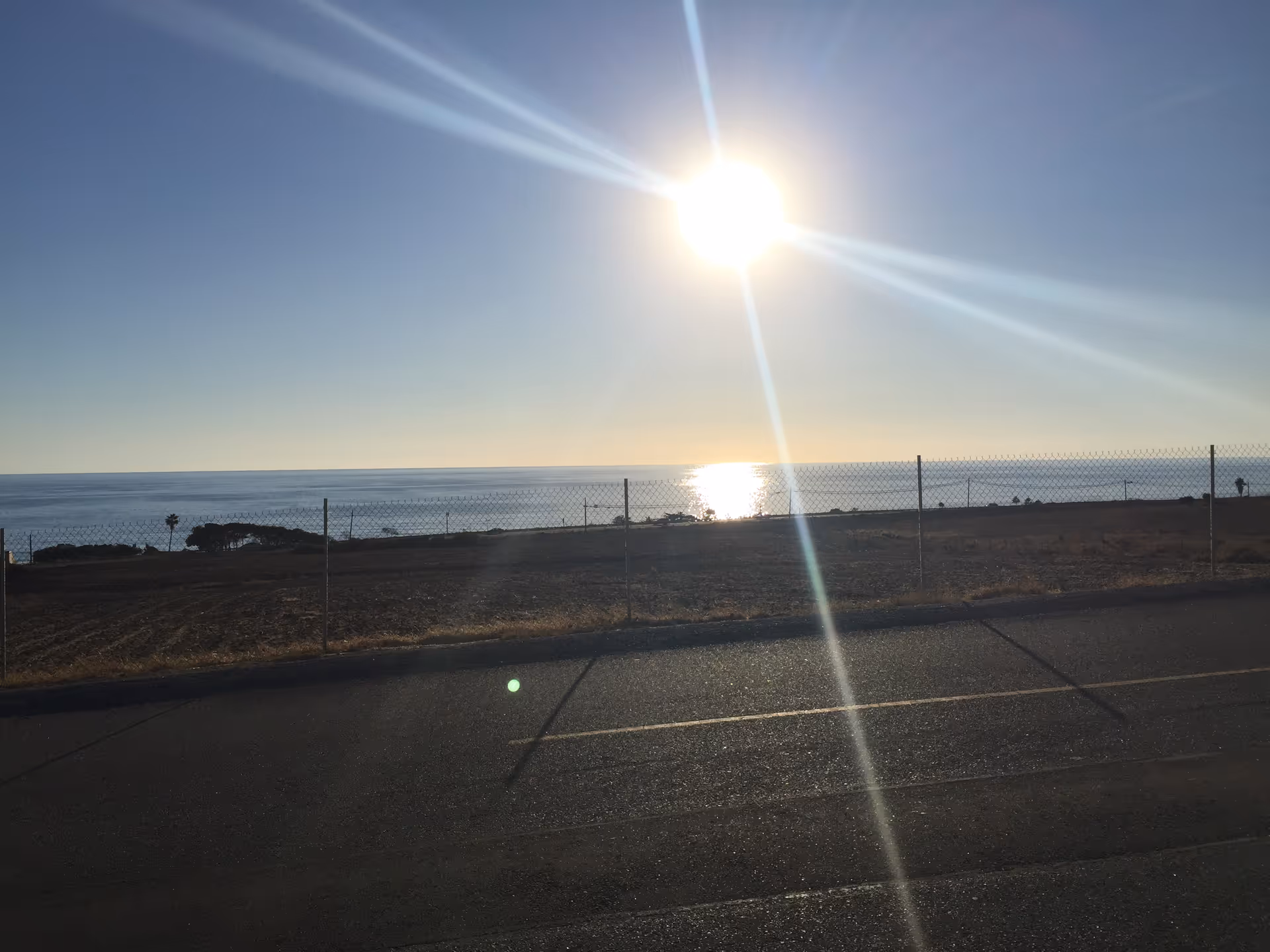 Sunny coastal scene with a fence, road, and calm ocean under bright sun
