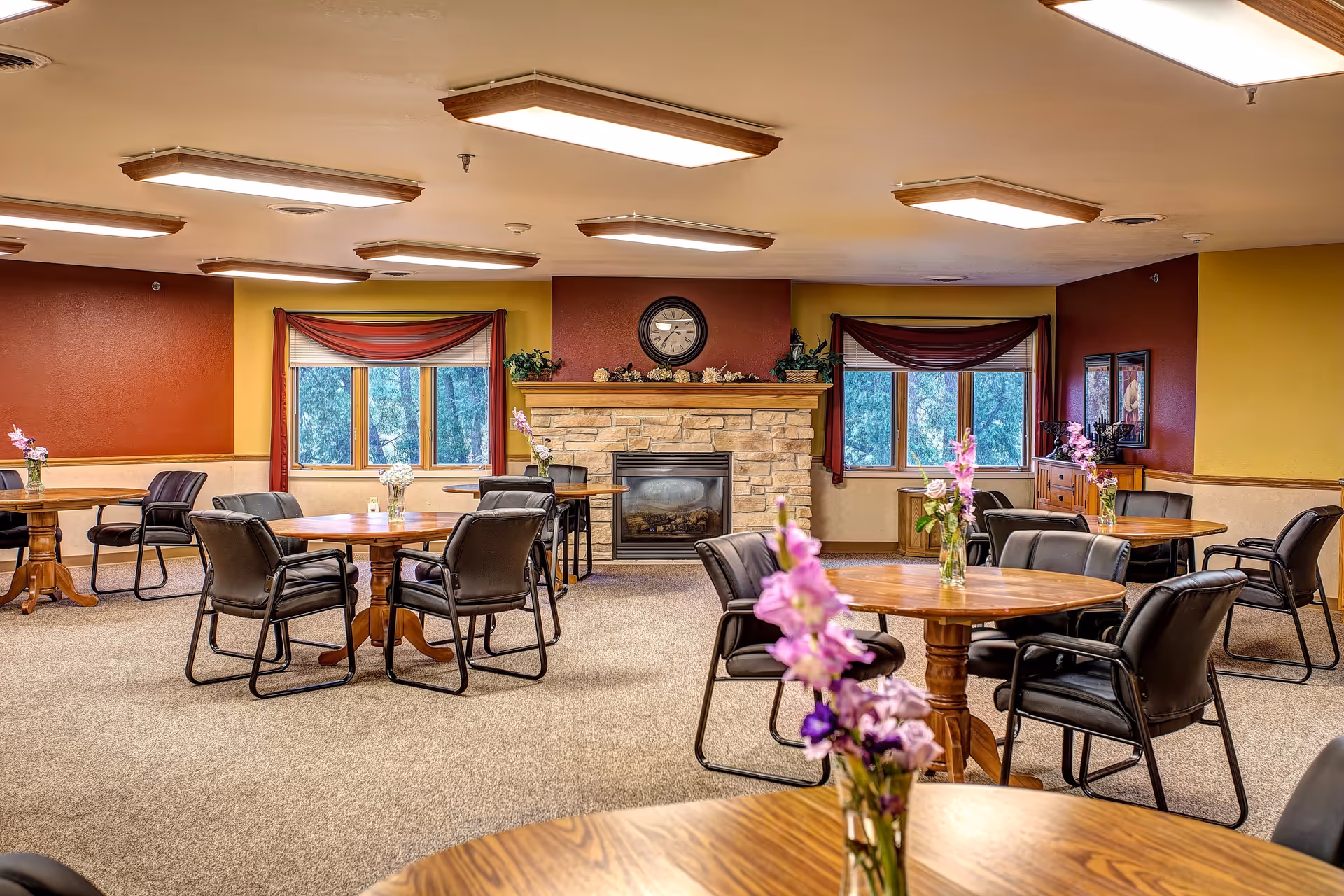 A spacious common area with multiple round wooden tables and black cushioned chairs arranged around them. Each table has a small vase with purple and white flowers. The room features a stone fireplace with a clock above it, flanked by two windows with red curtains. The walls are painted in warm tones of red and yellow, and the ceiling has several rectangular light fixtures.