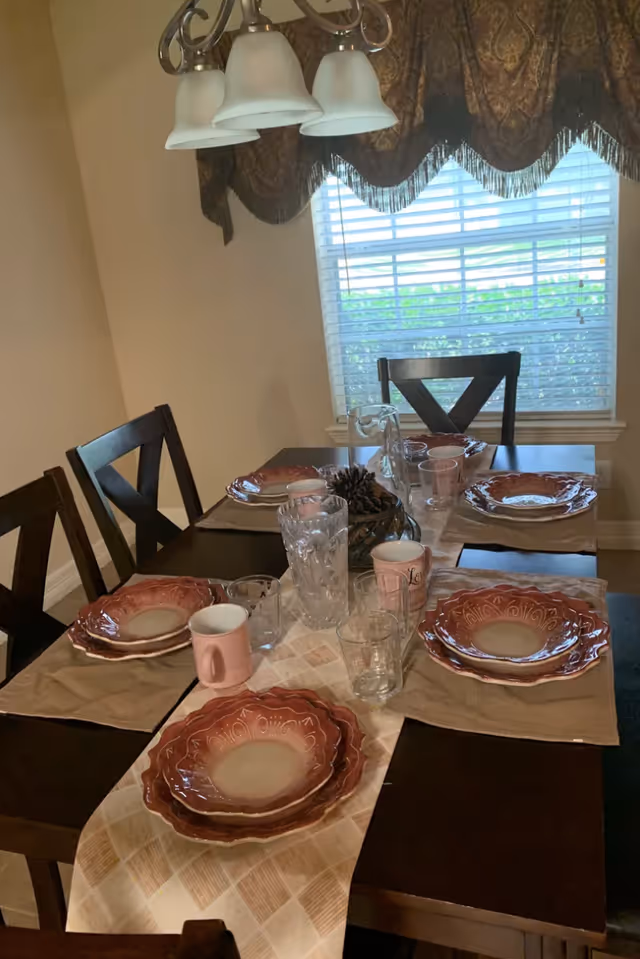Dining room table set for six with stacked red patterned plates, glasses, mugs, a pinecone centerpiece, and a hanging light fixture by a window with blinds.