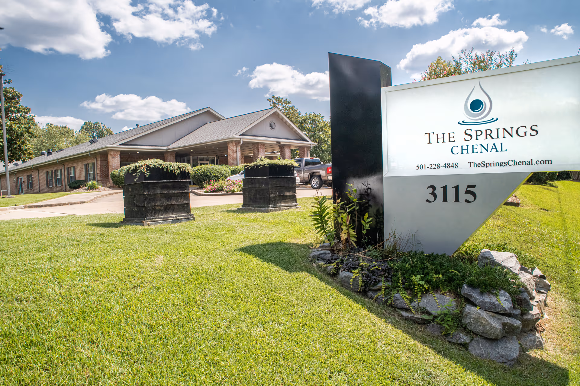 Exterior view of a senior living facility with sign, low brick building, and green lawn.