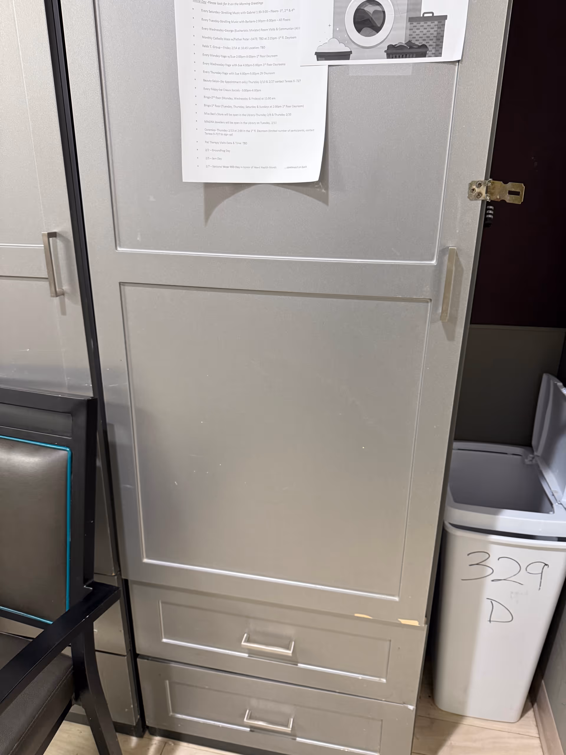 Tall gray cabinet with papers taped to front, trash can beside, chair in foreground.