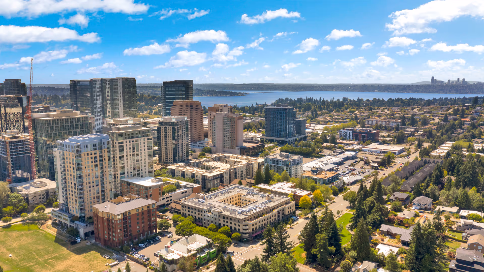 Aerial view of a cityscape featuring multiple mid-rise and high-rise buildings, residential areas, and a large body of water in the background under a partly cloudy blue sky.