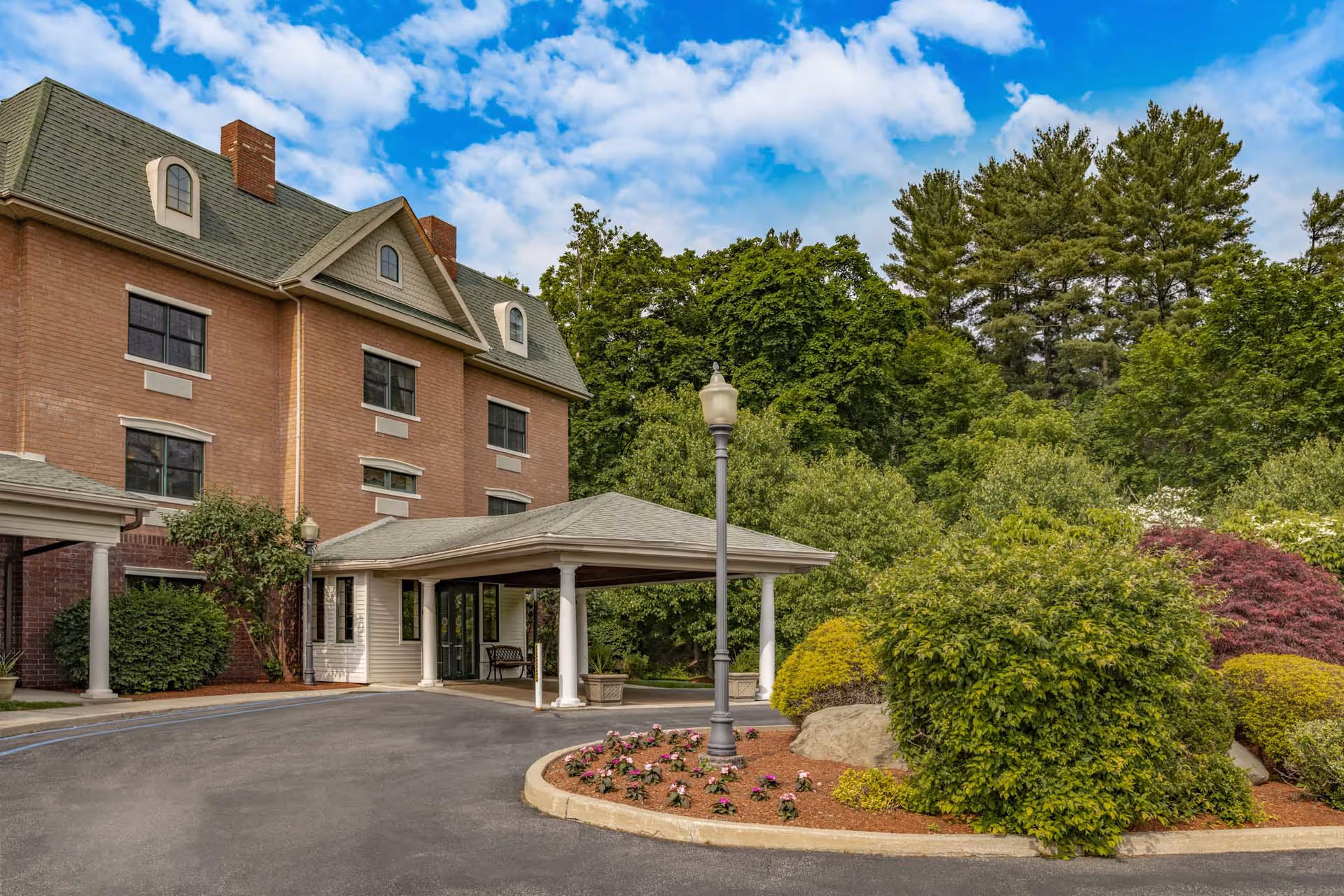 Exterior view of a senior living facility named Holiday Manor at Woodside, showing a multi-story brick building with a green roof, a covered entrance with white columns, a lamp post, and landscaped bushes and flowers under a partly cloudy blue sky.