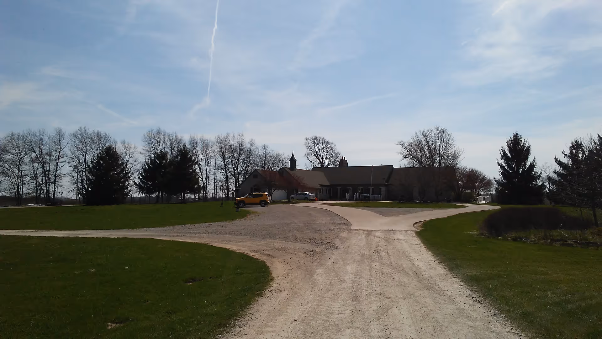 Gravel driveway curves toward a single-story facility with lawn and leafless trees.