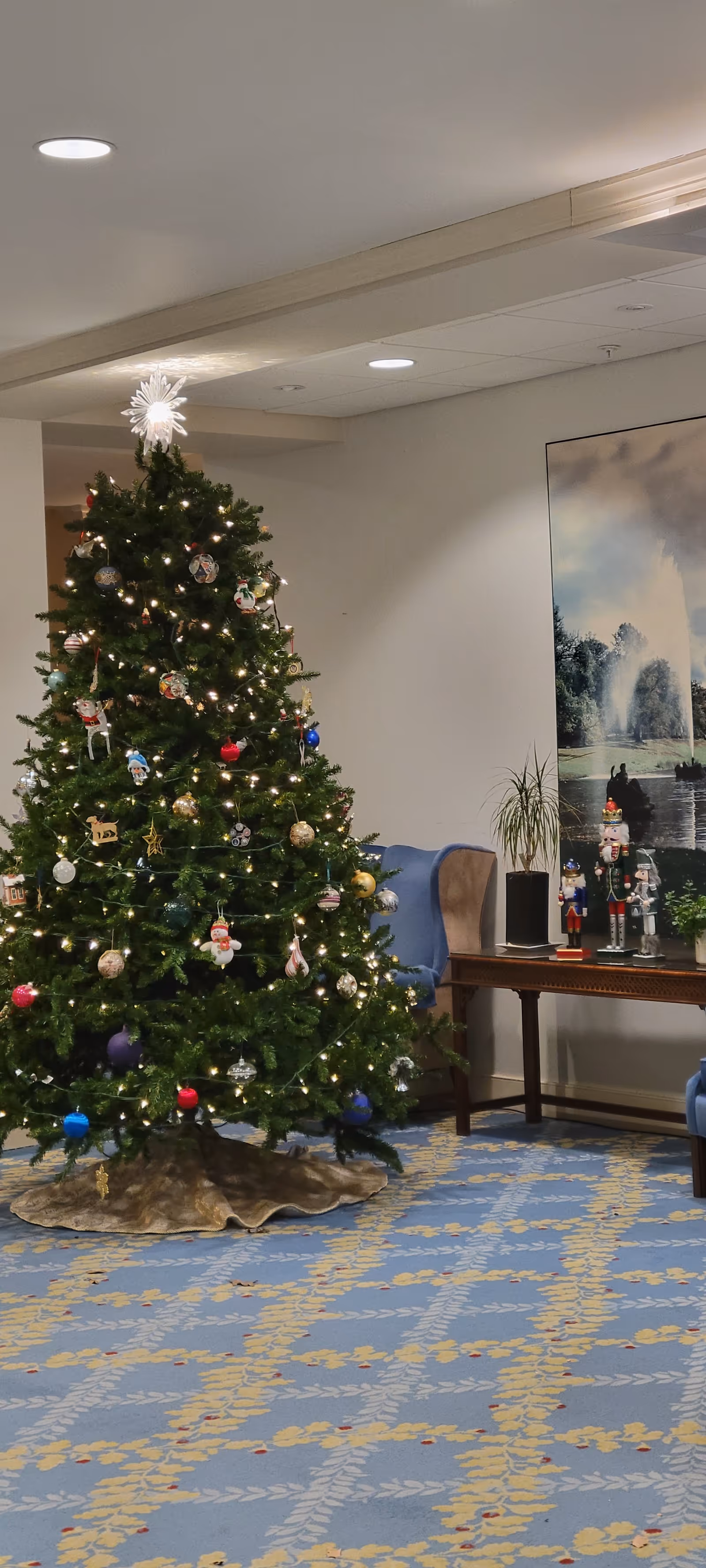 Decorated Christmas tree stands in a bright indoor lounge with chairs and artwork.