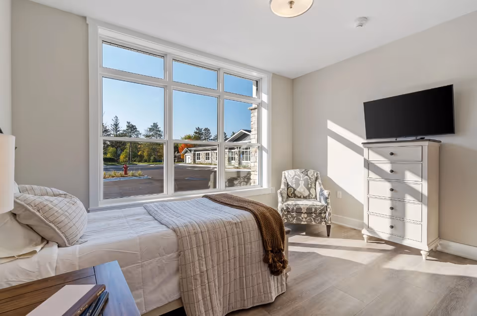 Bright bedroom with large window, bed, dresser, and TV in a senior living facility.