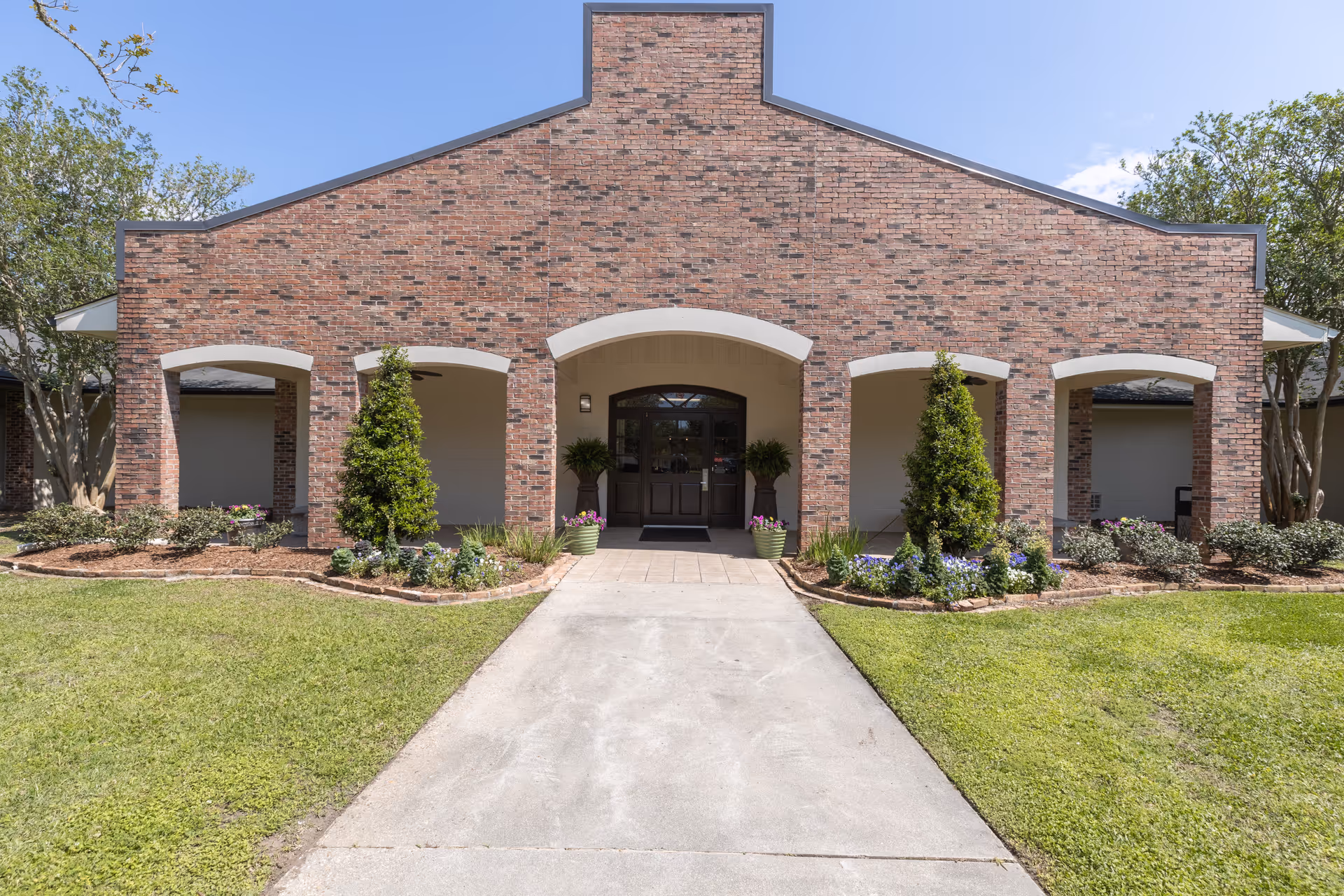 Front exterior view of a brick building with a central entrance featuring a dark door and two potted plants on either side. The building has four arches supported by brick columns, with landscaped flower beds and small trees in front. A concrete walkway leads up to the entrance, and the sky is clear and blue.