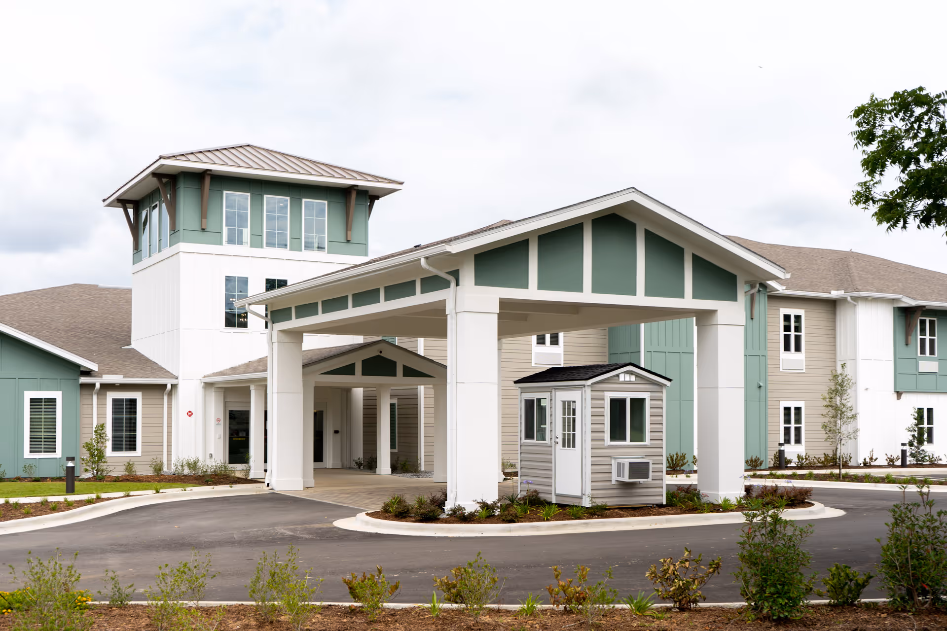 Exterior view of The Canopy at Azalea Grove senior living facility showing a covered entrance with a small guardhouse, surrounded by landscaped greenery and a paved driveway.
