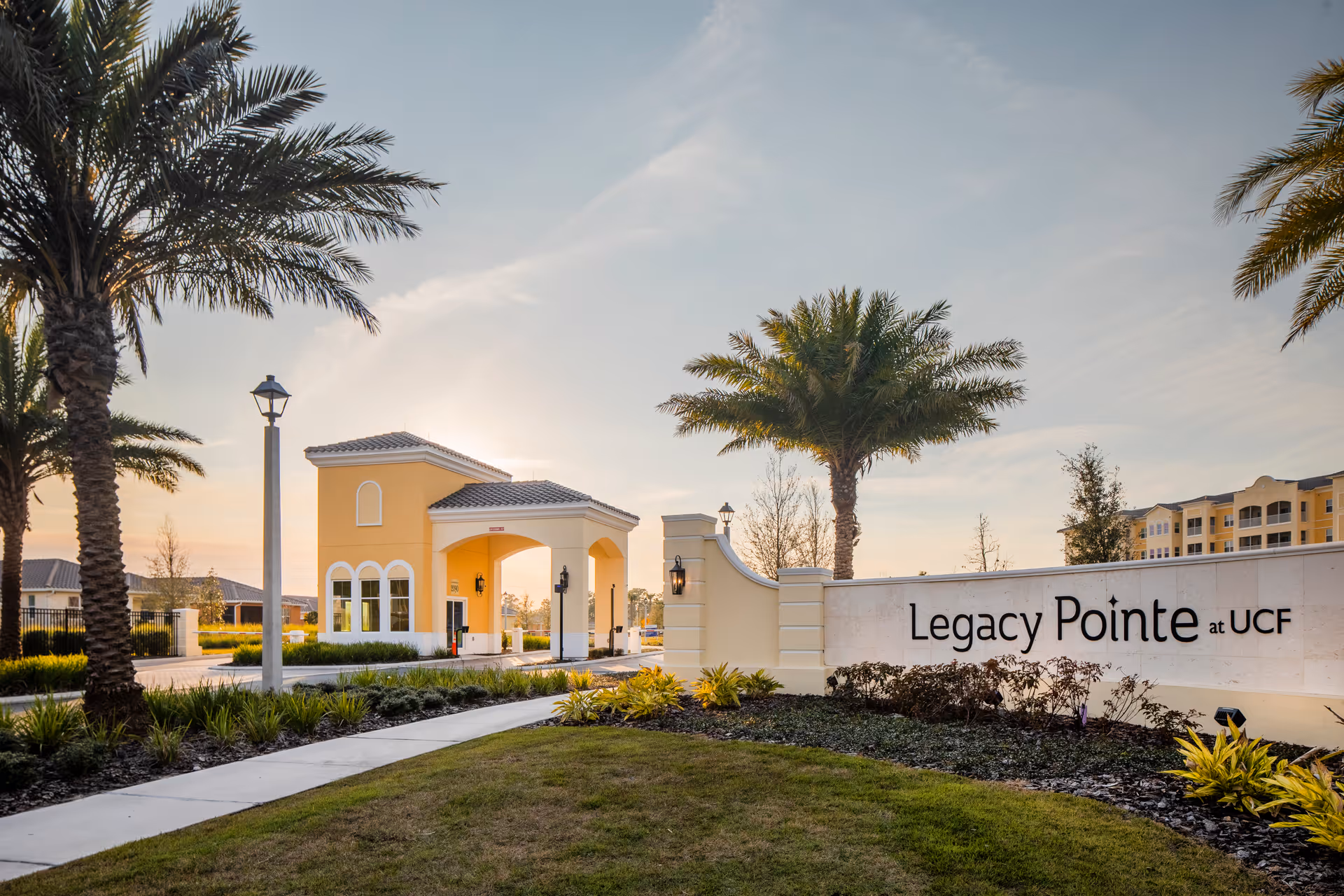 Entrance gate and landscaped sign reading 'Legacy Pointe at UCF' with a guardhouse, palm trees, and sunset sky.