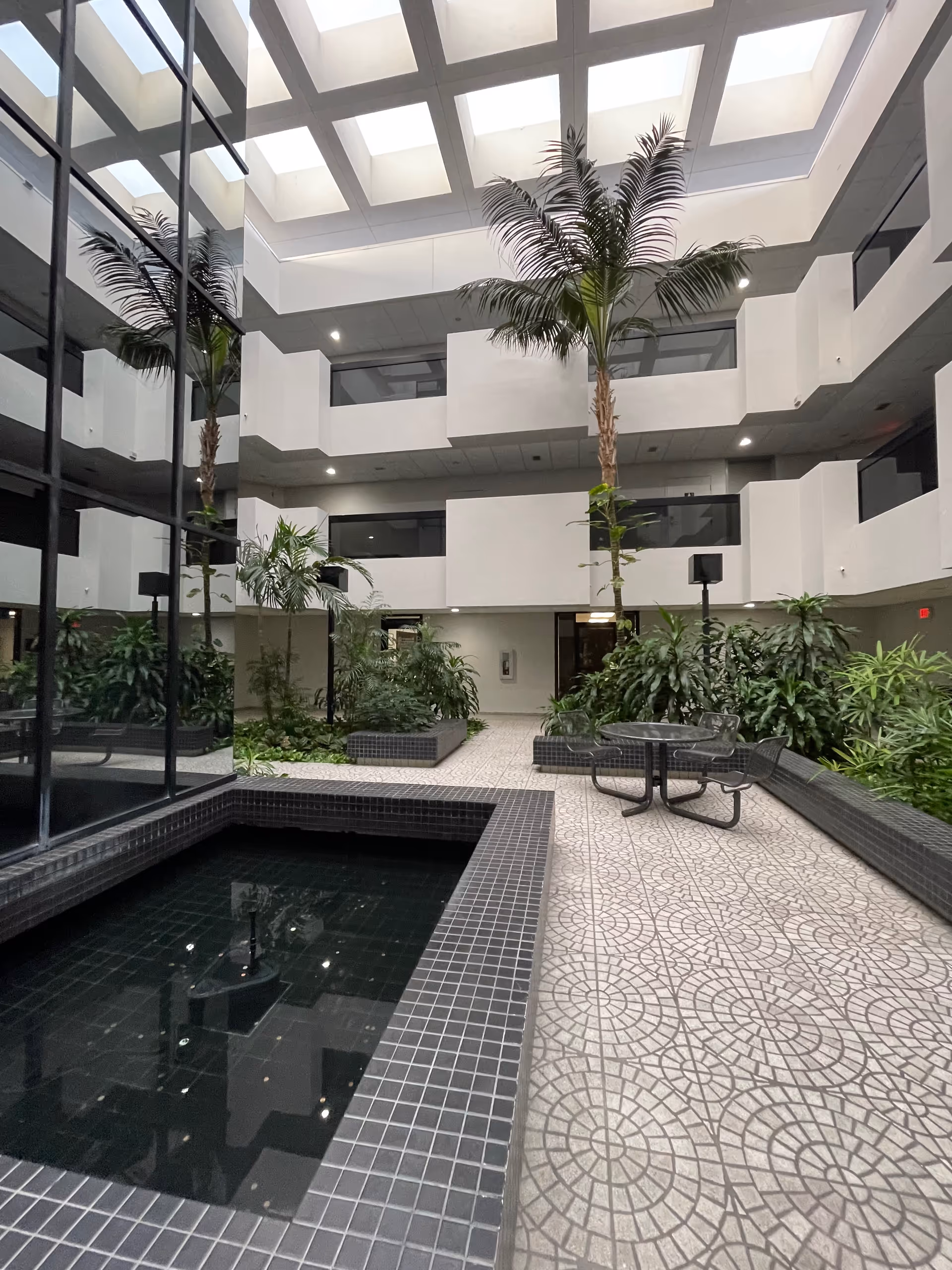 Indoor atrium with skylights, tall palm trees, planters, and a tiled water feature near seating.