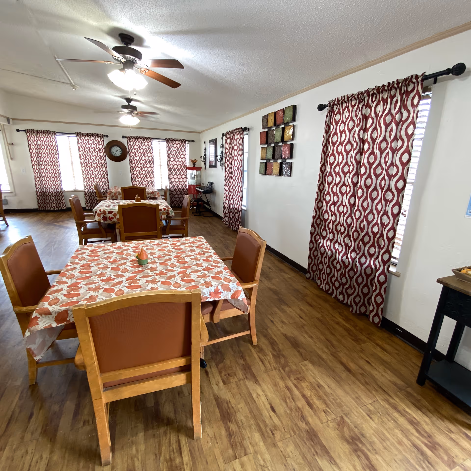 A dining room with wooden floors and several tables covered with autumn-themed tablecloths. Each table is surrounded by wooden chairs with brown cushions. The room has multiple windows with red and white patterned curtains, two ceiling fans with lights, a wall clock, and decorative wall art. There is also a small popcorn machine and a keyboard in the corner.
