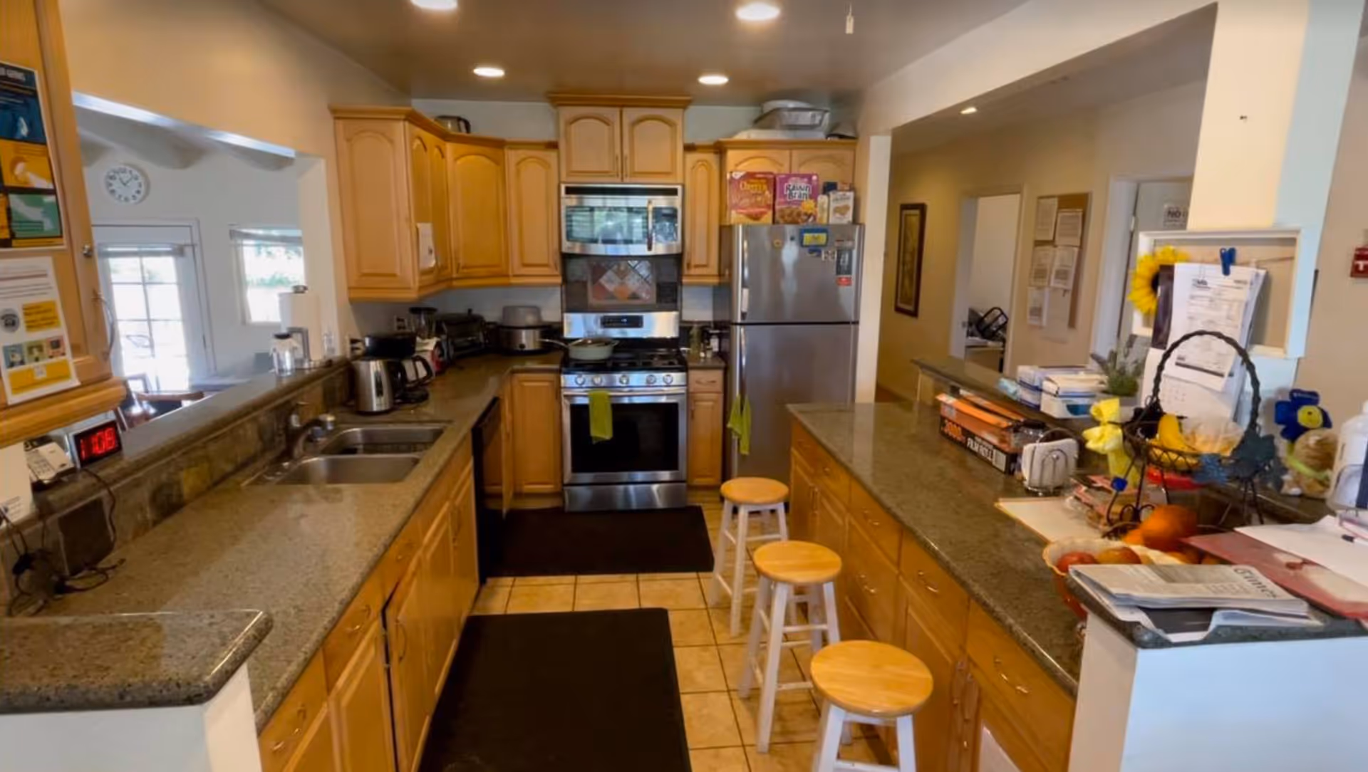 A well-lit kitchen with wooden cabinets, granite countertops, and tiled floor. The kitchen features a stainless steel stove and refrigerator, a double sink, and various small appliances on the counters. There are three wooden stools along the kitchen island, and some cereal boxes on top of the refrigerator. The kitchen opens into a hallway and another room with windows.