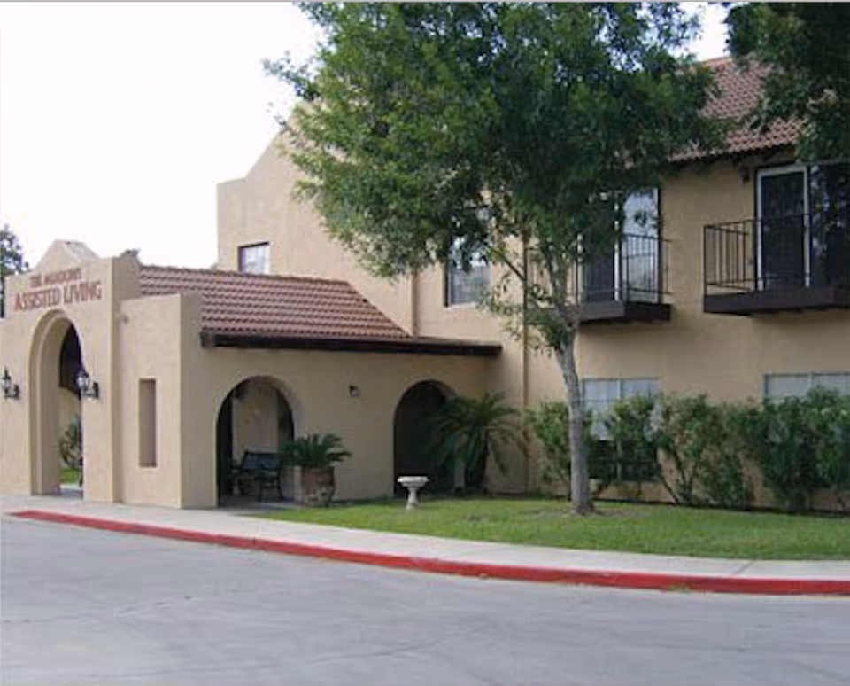 Exterior view of Spanish Meadows of Brownsville assisted living facility showing a beige building with a red-tiled roof, arched entryway, balconies, trees, and a small lawn area with shrubs and a birdbath.