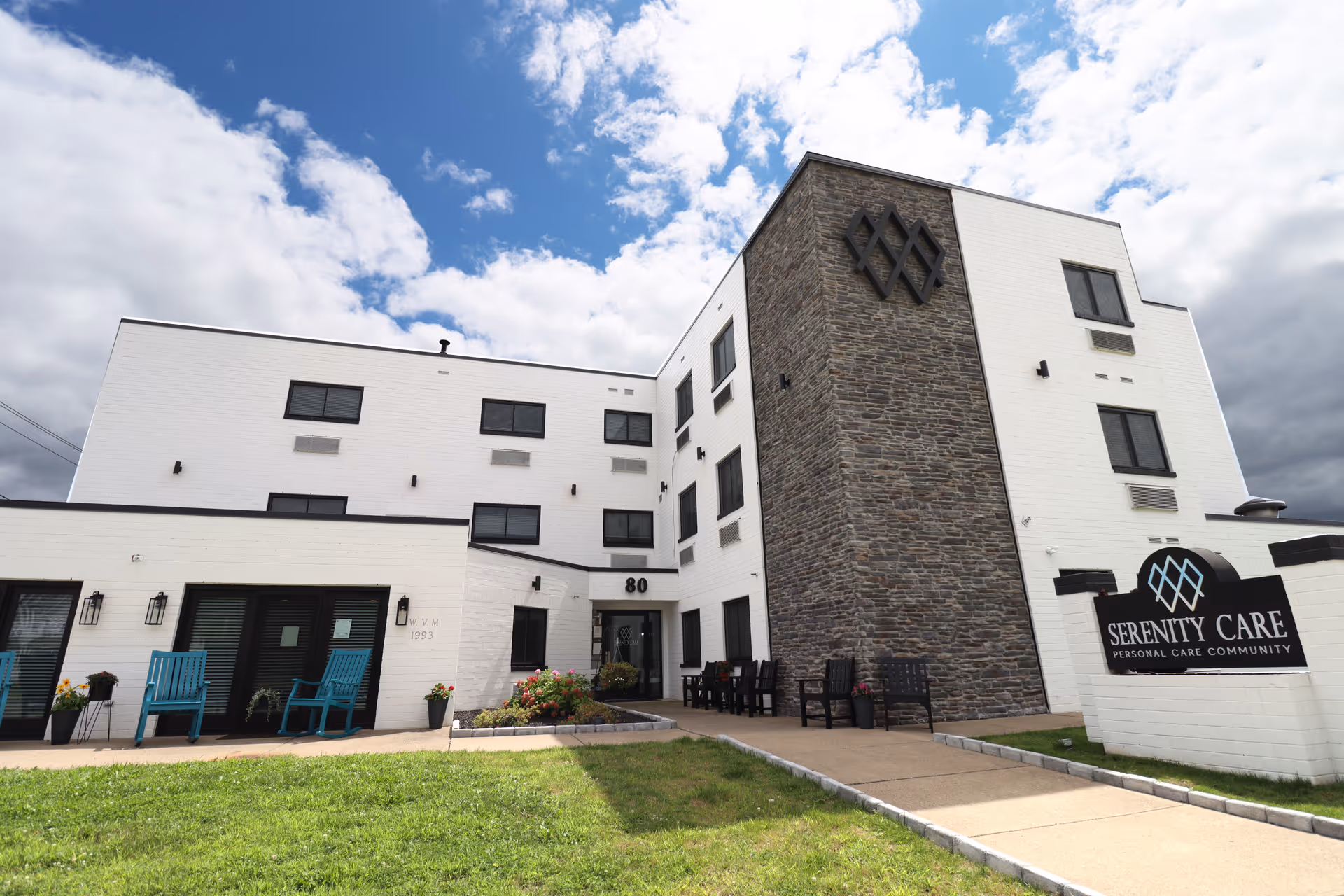 Exterior view of a modern three-story building with white brick walls and a stone accent wall. The building has several windows and a main entrance with the number 80 above the door. There are blue chairs and potted plants near the entrance, and a sign in front reads 'Serenity Care Personal Care Community'. The sky is partly cloudy.