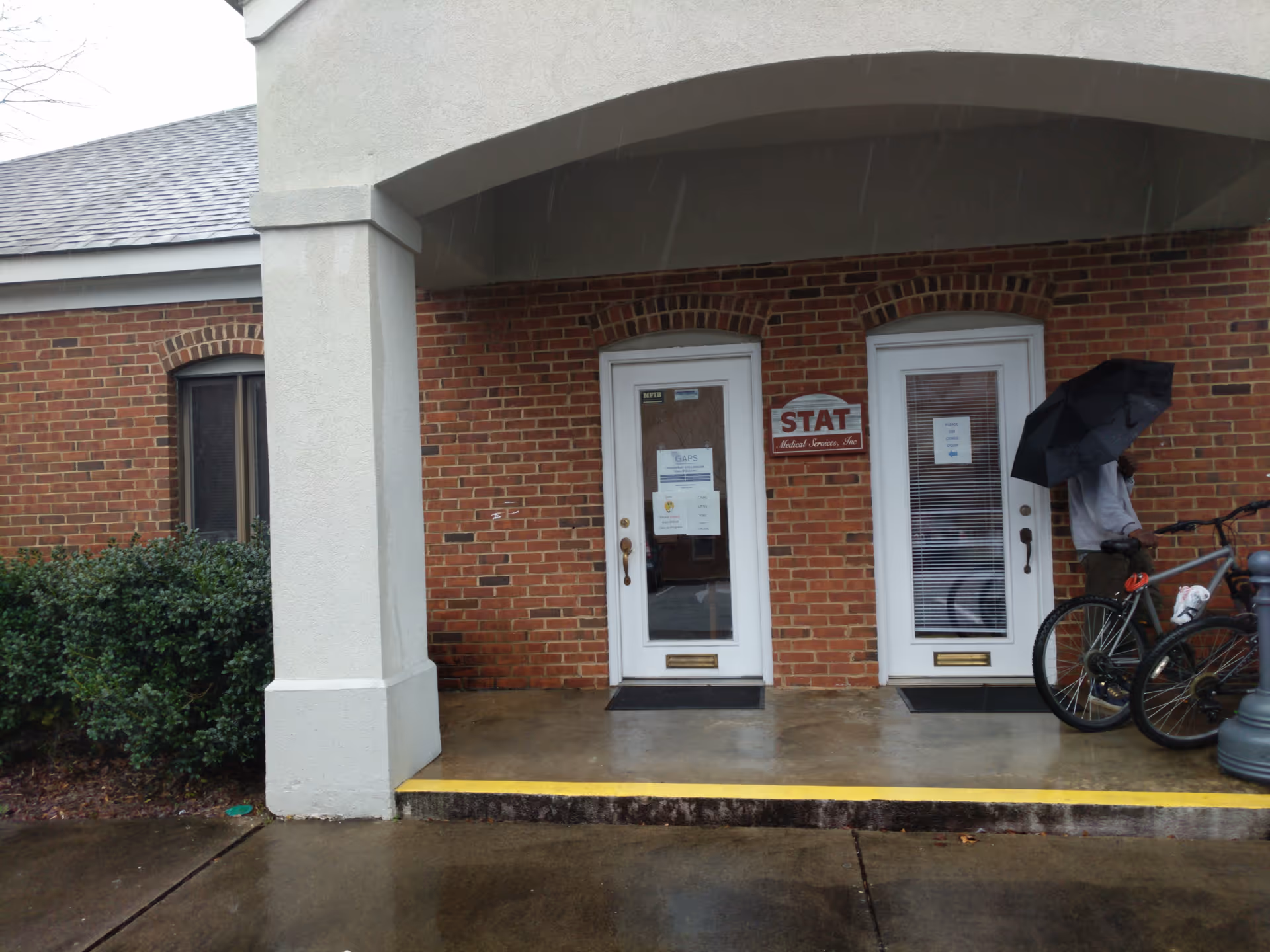 Brick building entrance with two doors labeled STAT, bikes, and a person with umbrella outside.
