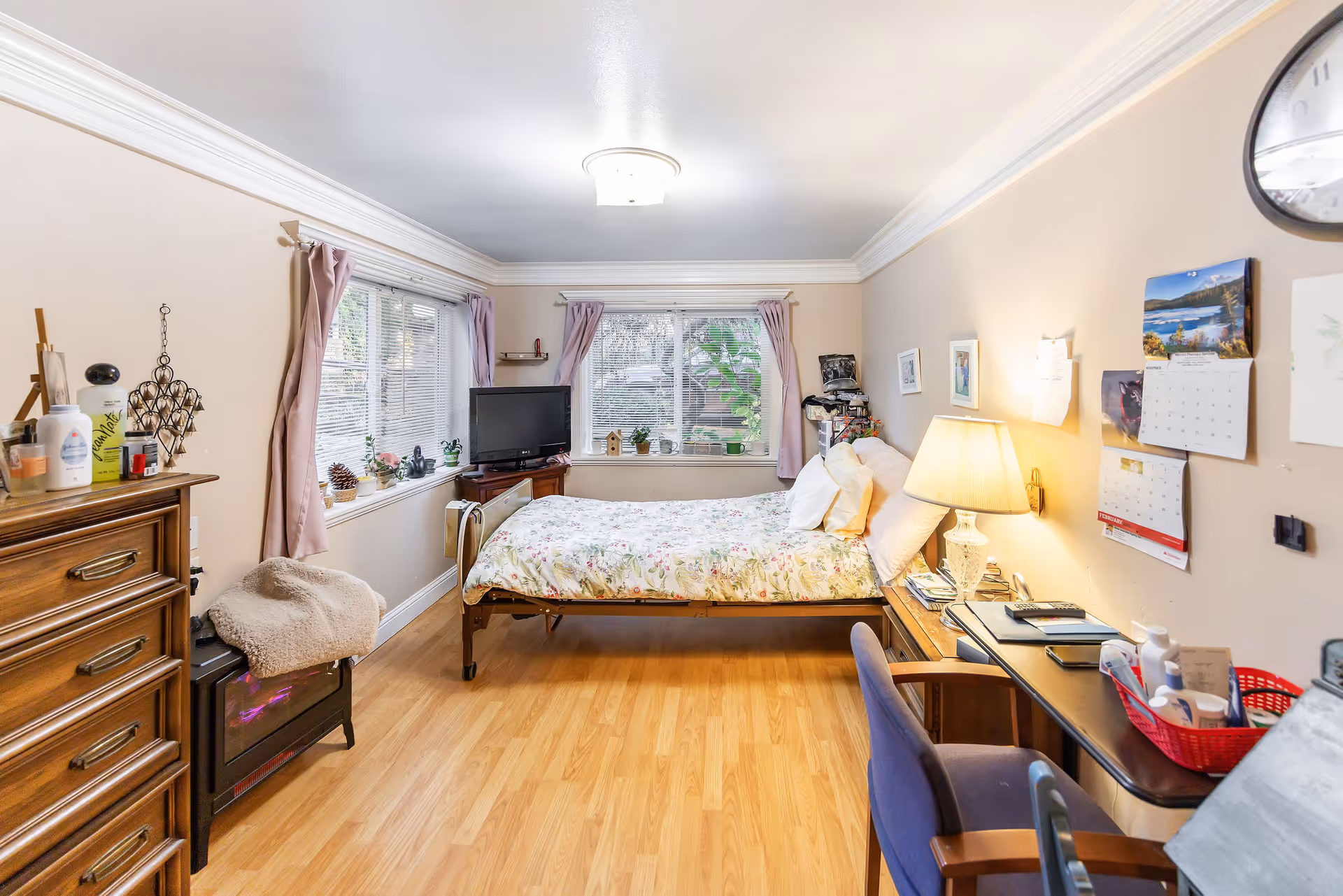 Cozy bedroom in a senior living facility with a bed, dresser, desk, and windows.