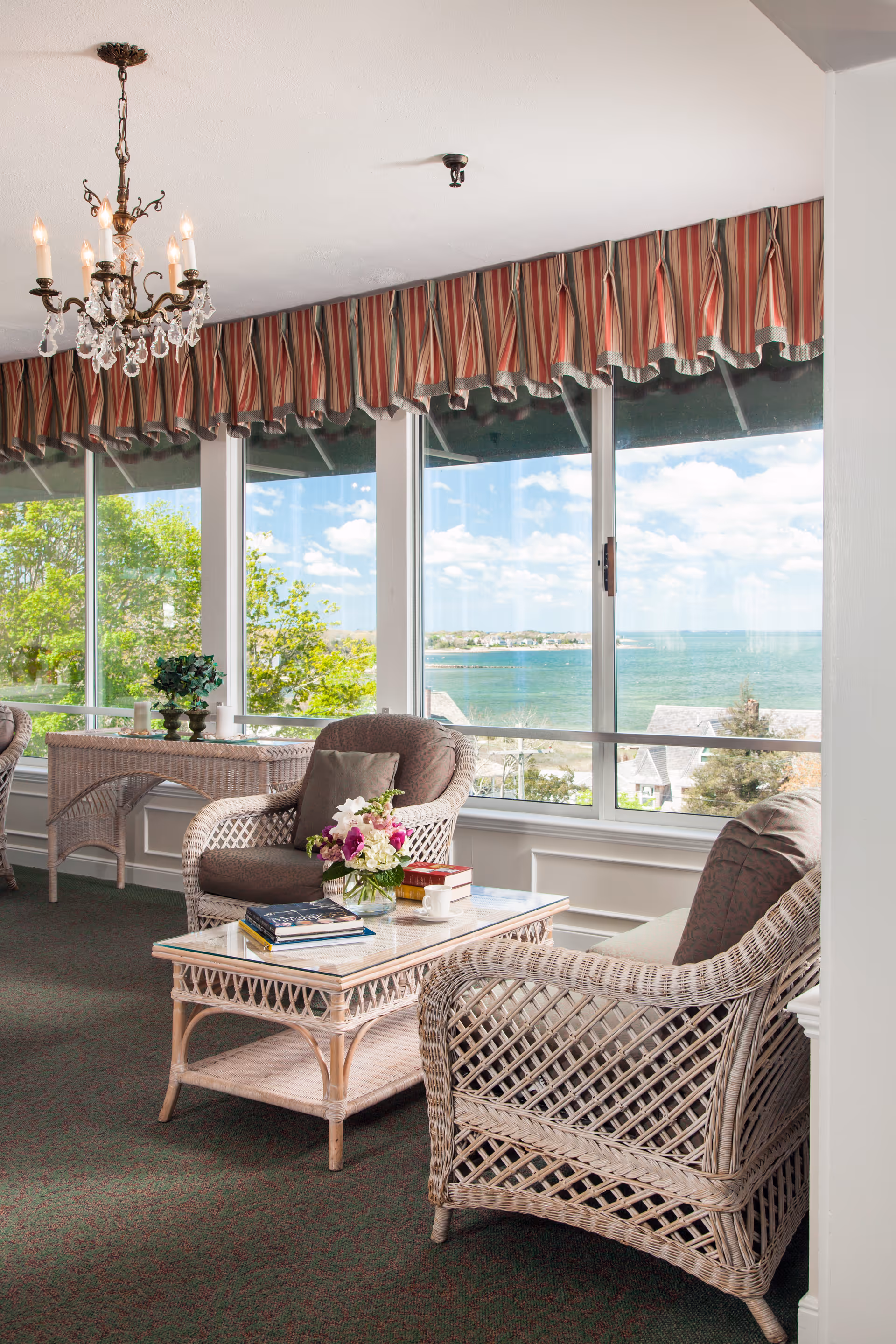 Sunlit sitting area with wicker armchairs, a glass-top coffee table, and large windows overlooking a waterfront view.